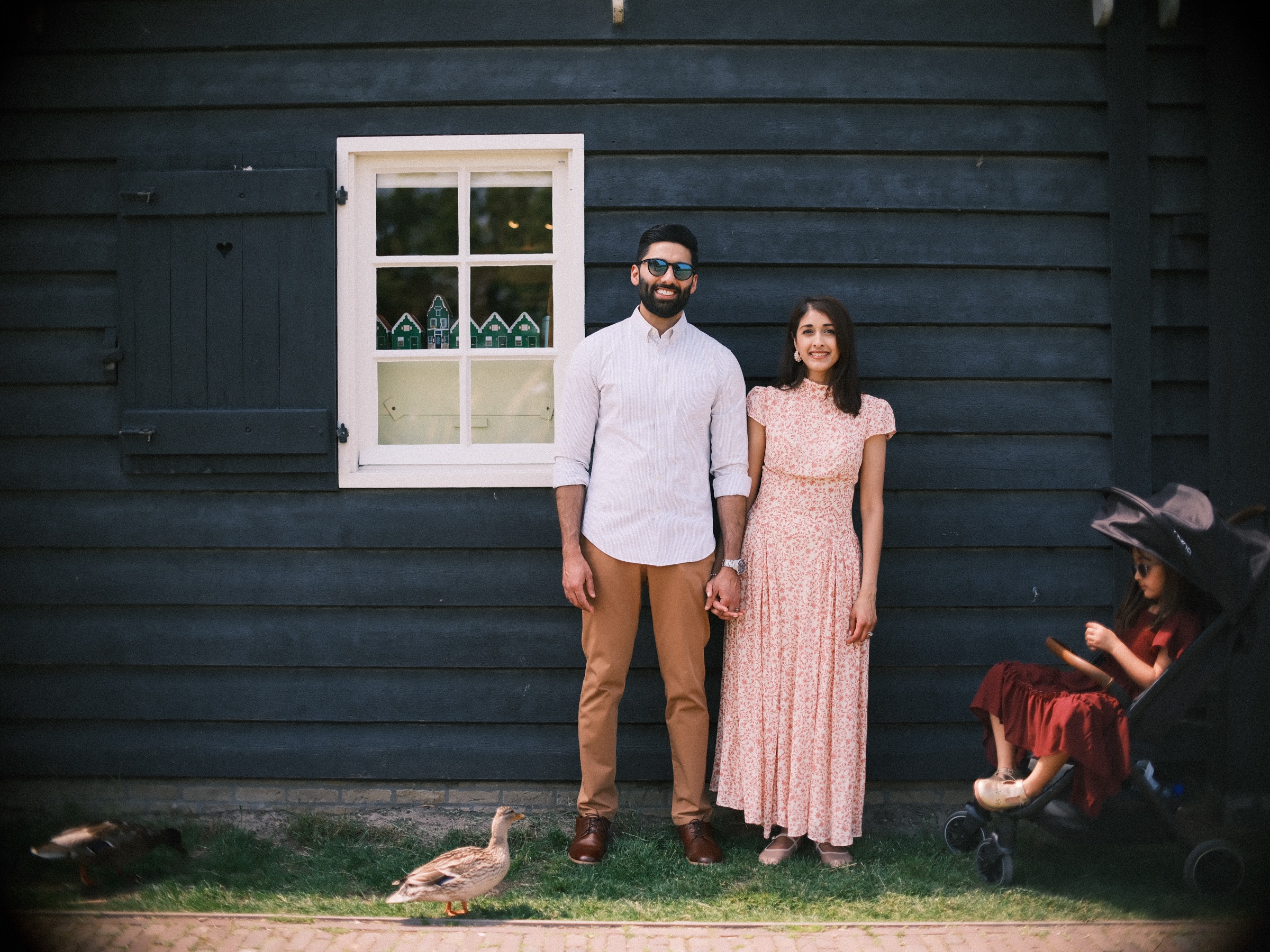 Parents holding hands by a dark wooden house while their daughter rests in a stroller and a duck walks past at the Zaanse Schans