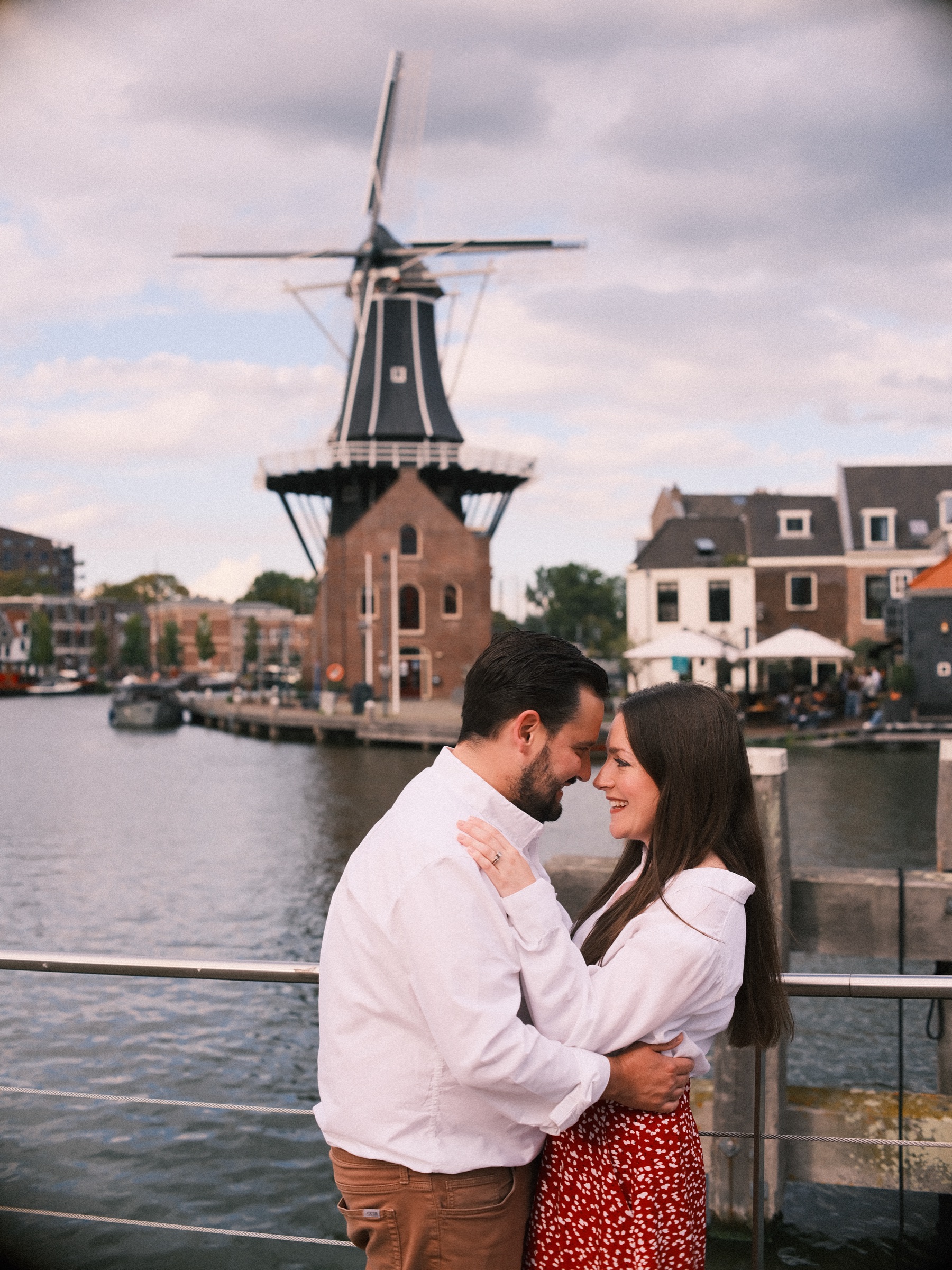 Man in een wit overhemd en vrouw in een rode rok staan voorhoofd tegen voorhoofd bij de reling, met de molen en het water erachter