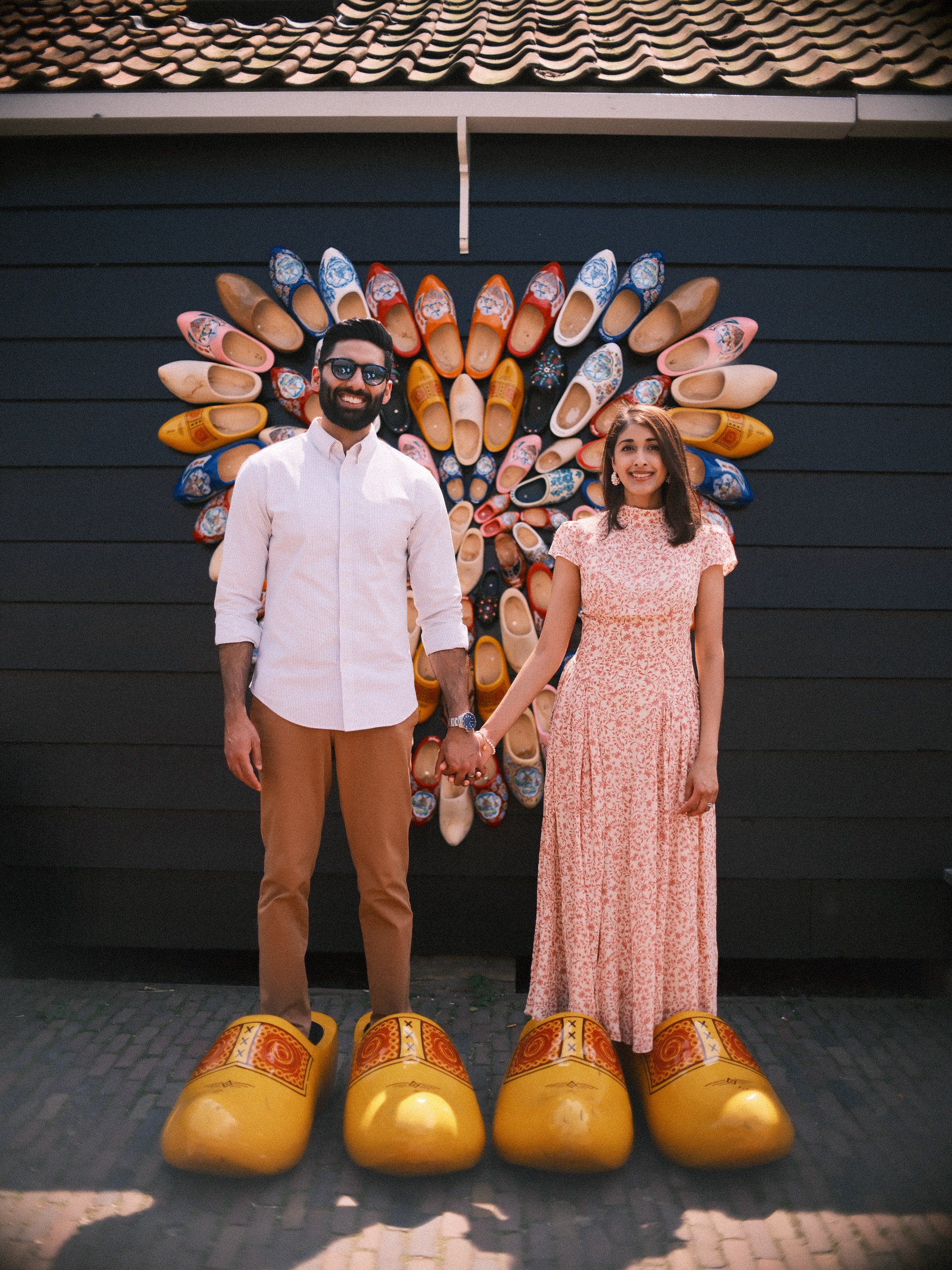 Couple holding hands before a heart of painted wooden clogs on a dark wall at the Zaanse Schans, giant yellow clogs at their feet