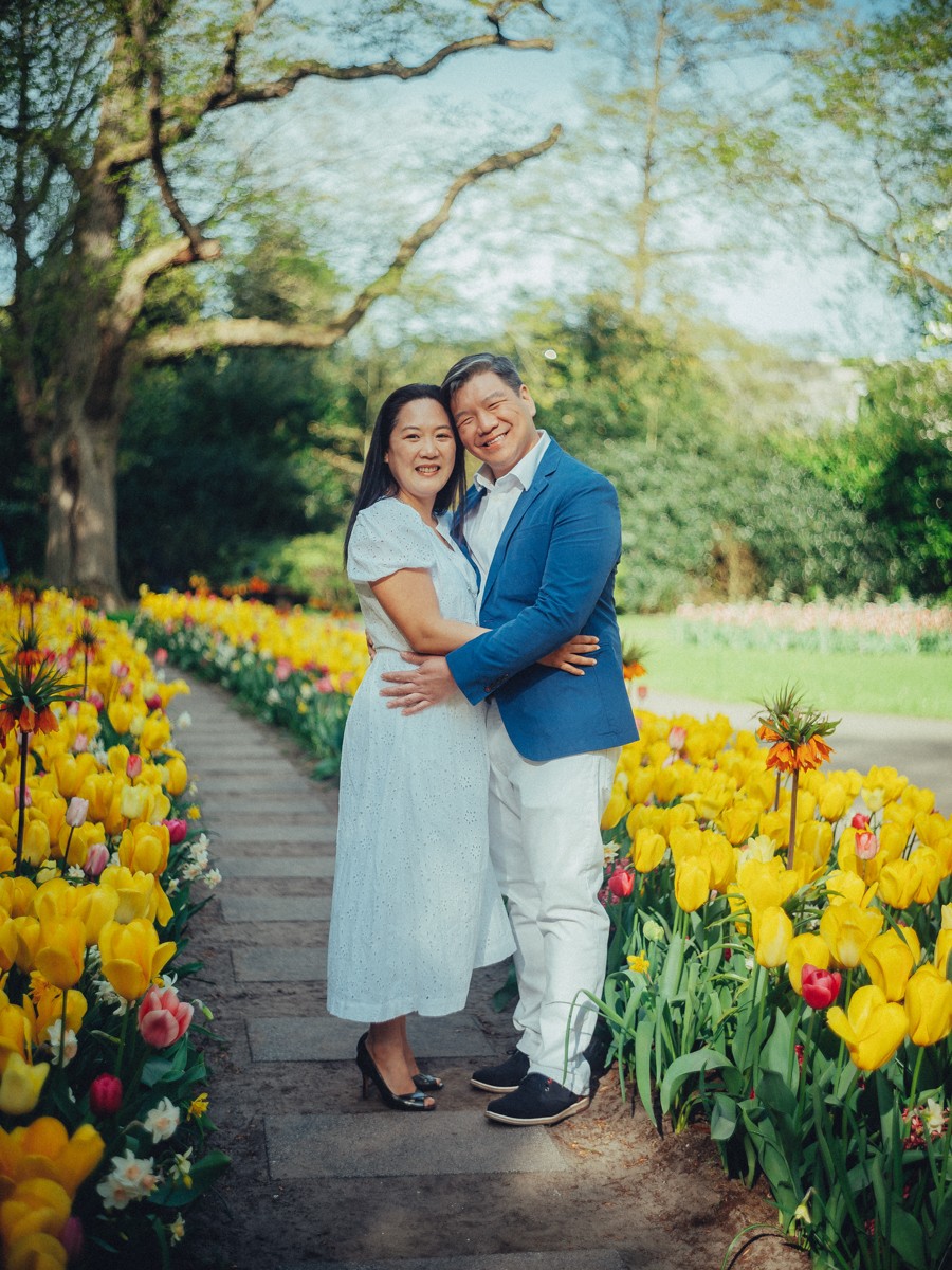 Vader in een blauw colbert houdt moeder dicht tegen zich aan op een stenen pad tussen gele tulpen en keizerskronen