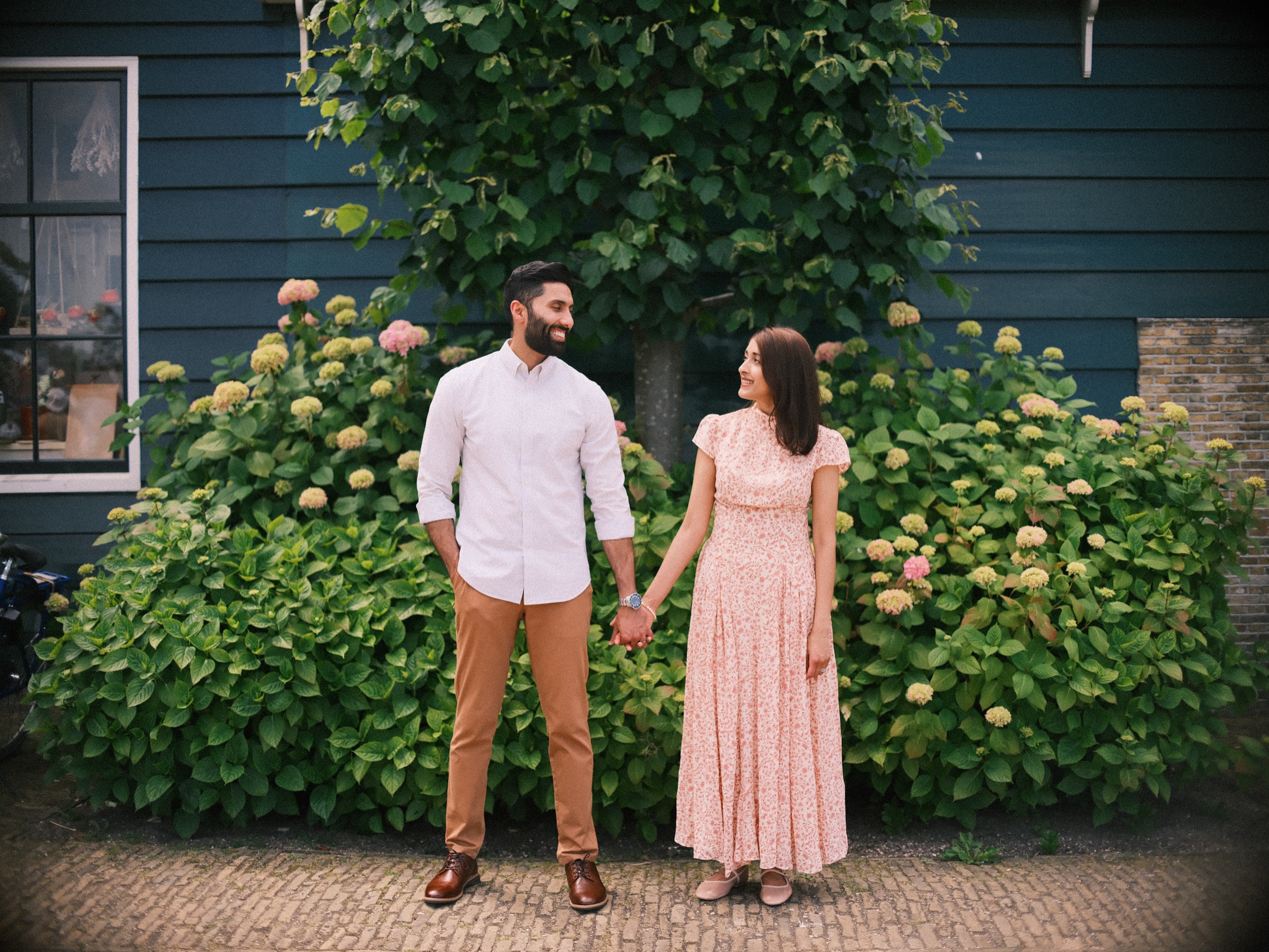Parents holding hands in front of a large hydrangea bush against a dark blue wooden house, he in a white shirt, she in a pink floral dress