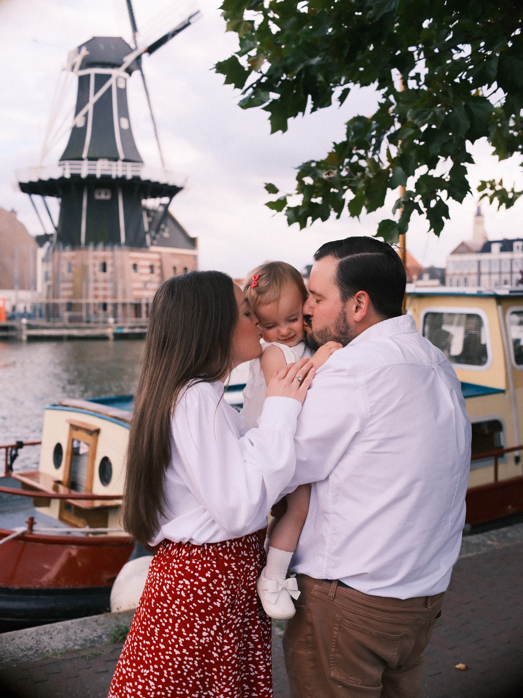 Ouders van achteren gezien kussen hun dochtertje van twee kanten op haar wangen, met een bootje en de molen voor hen