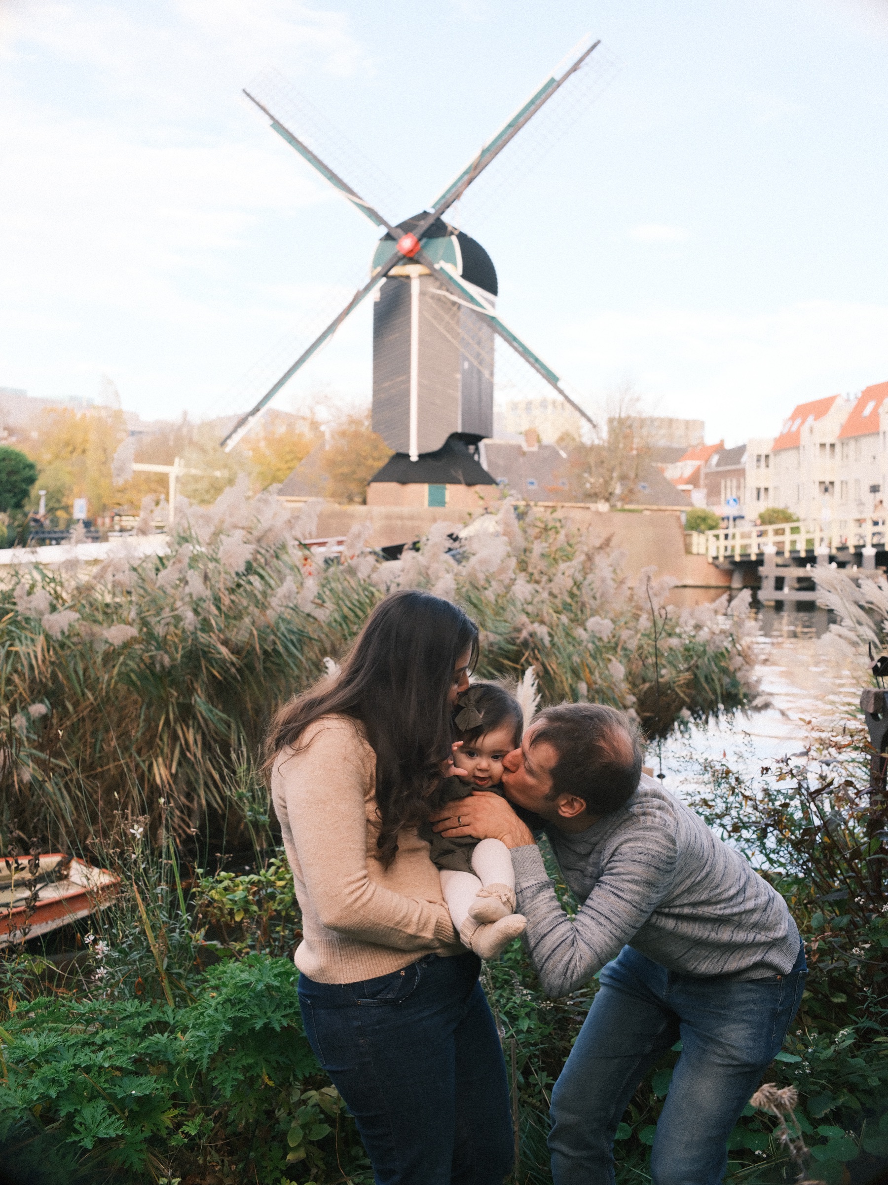 Parents kissing their baby's cheeks at the same time among tall autumn grasses, Molen De Put across the water behind them