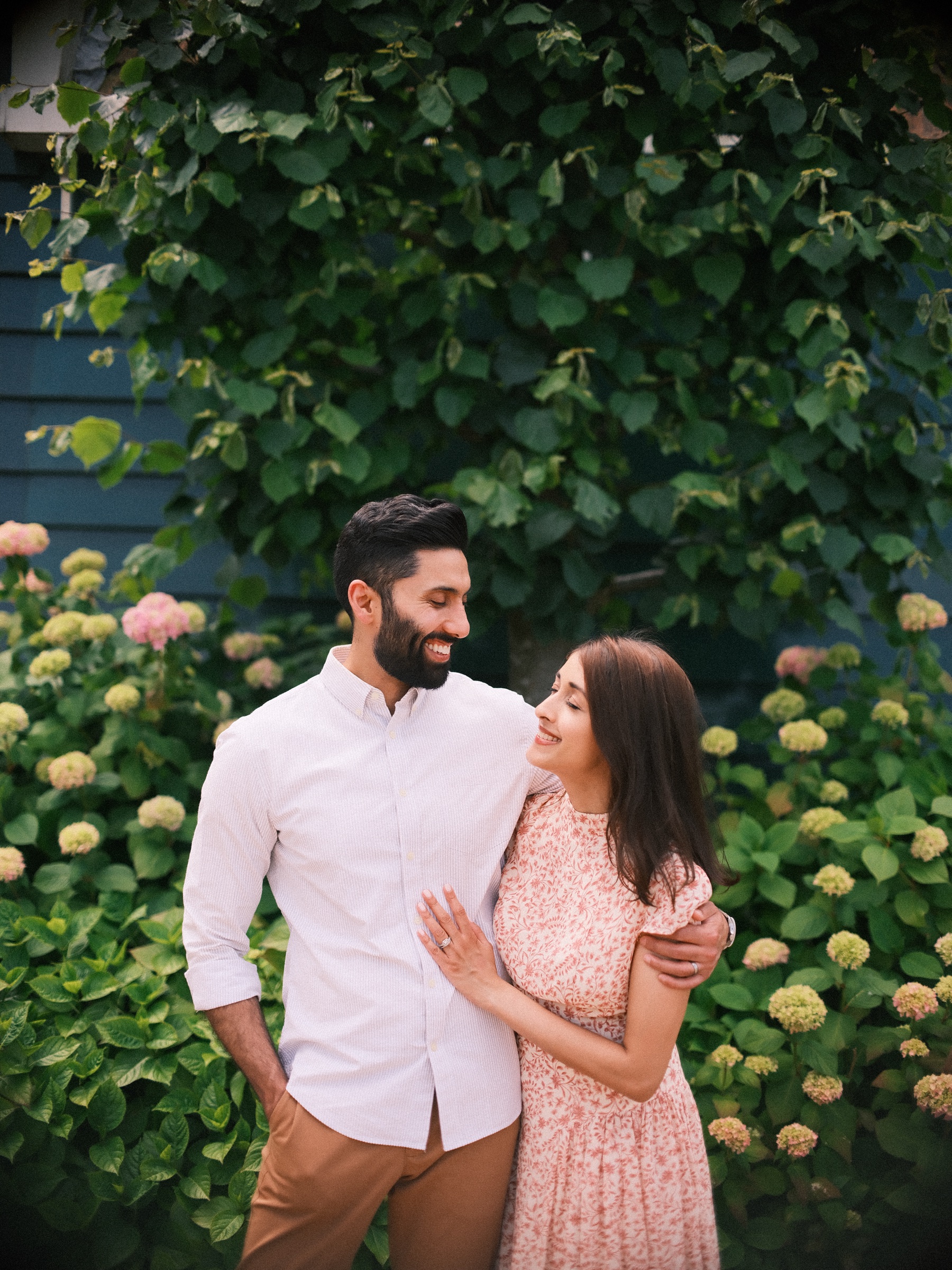 Mother in a pink floral maxi dress smiling up at her husband in a white shirt as they stand close in front of green hydrangeas