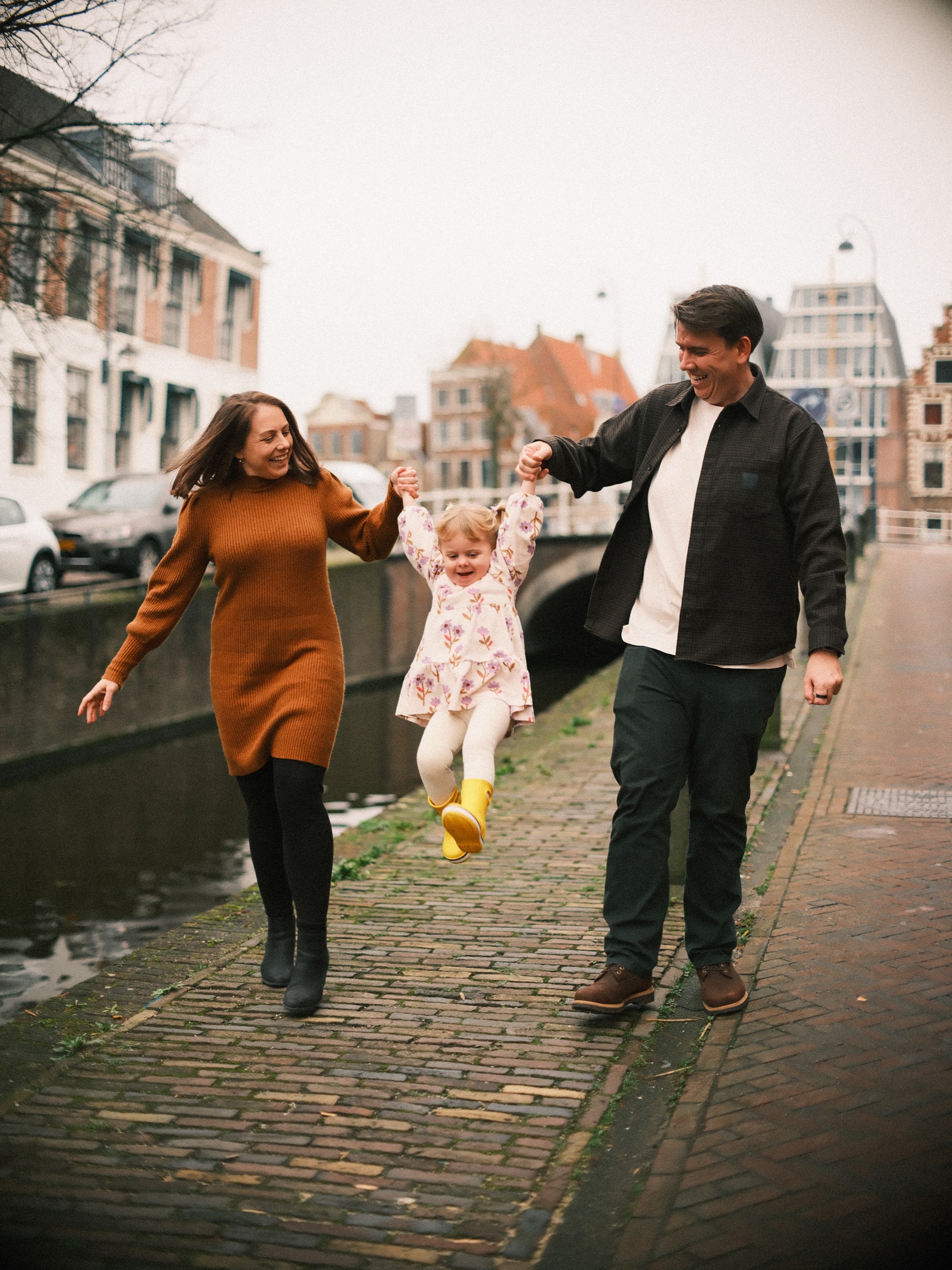 Ouders zwaaien hun peuter aan haar handen tussen zich in langs een gracht in Haarlem, met een trapgevel en een wit gebouw erachter