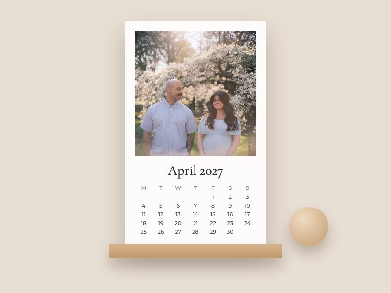 Desk calendar for April 2027 on a wooden stand, with a couple expecting a baby under white blossoms, beside a wooden ball