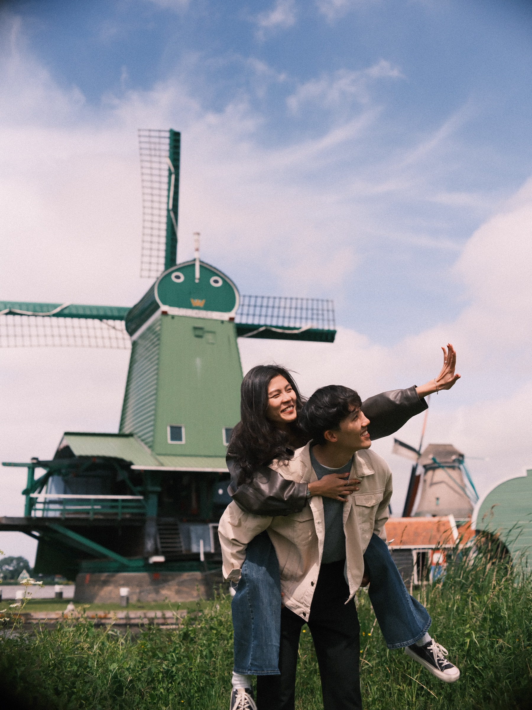 Laughing woman riding piggyback on her fiancé with one arm stretched toward the sky, the green mill and a gabled house behind them