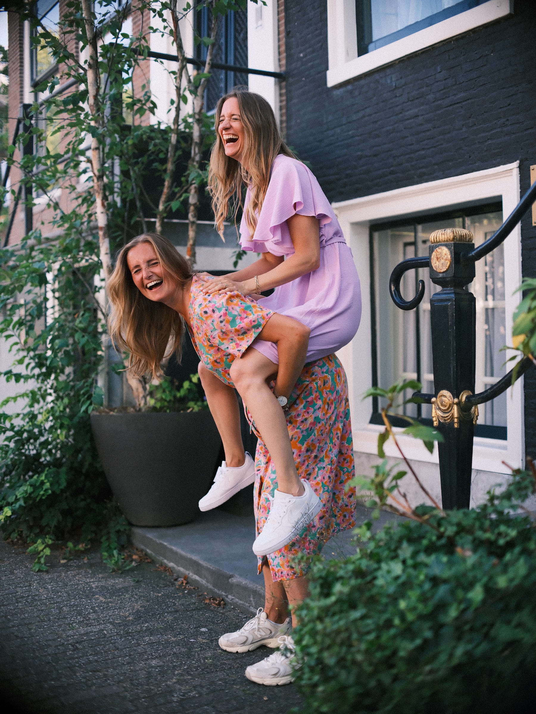 Bride-to-be riding piggyback on her best friend's back by a canal house, both laughing, at her bachelorette with friends