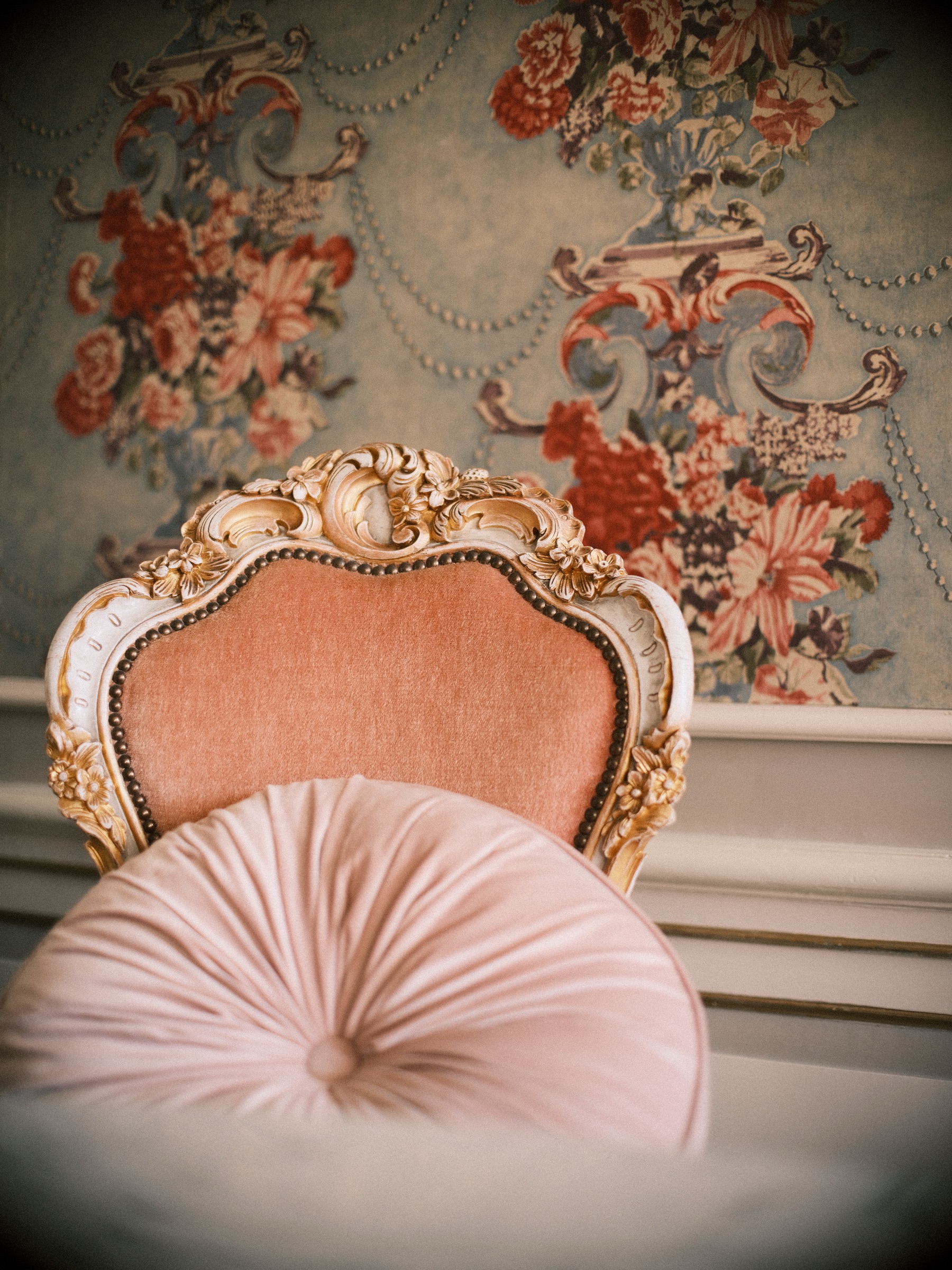 Roze fluwelen stoel met een bewerkte vergulde lijst en een rond kussen, met zacht onscherp bloemenbehang erachter