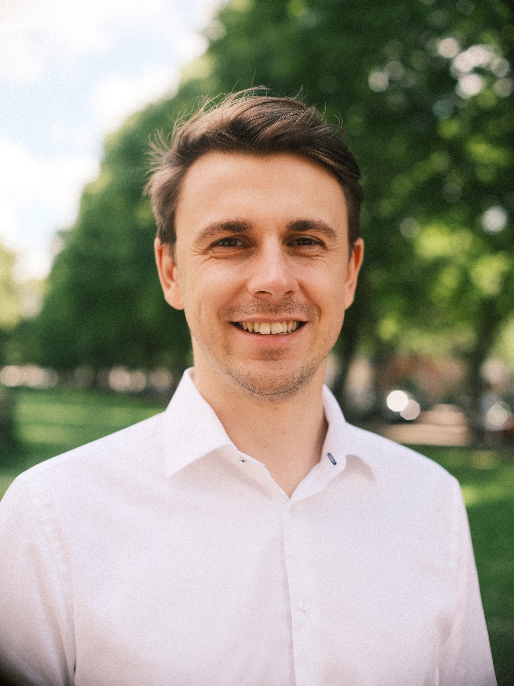 Smiling portrait of him in a white shirt against soft green trees, a single frame at the end of their engagement photos