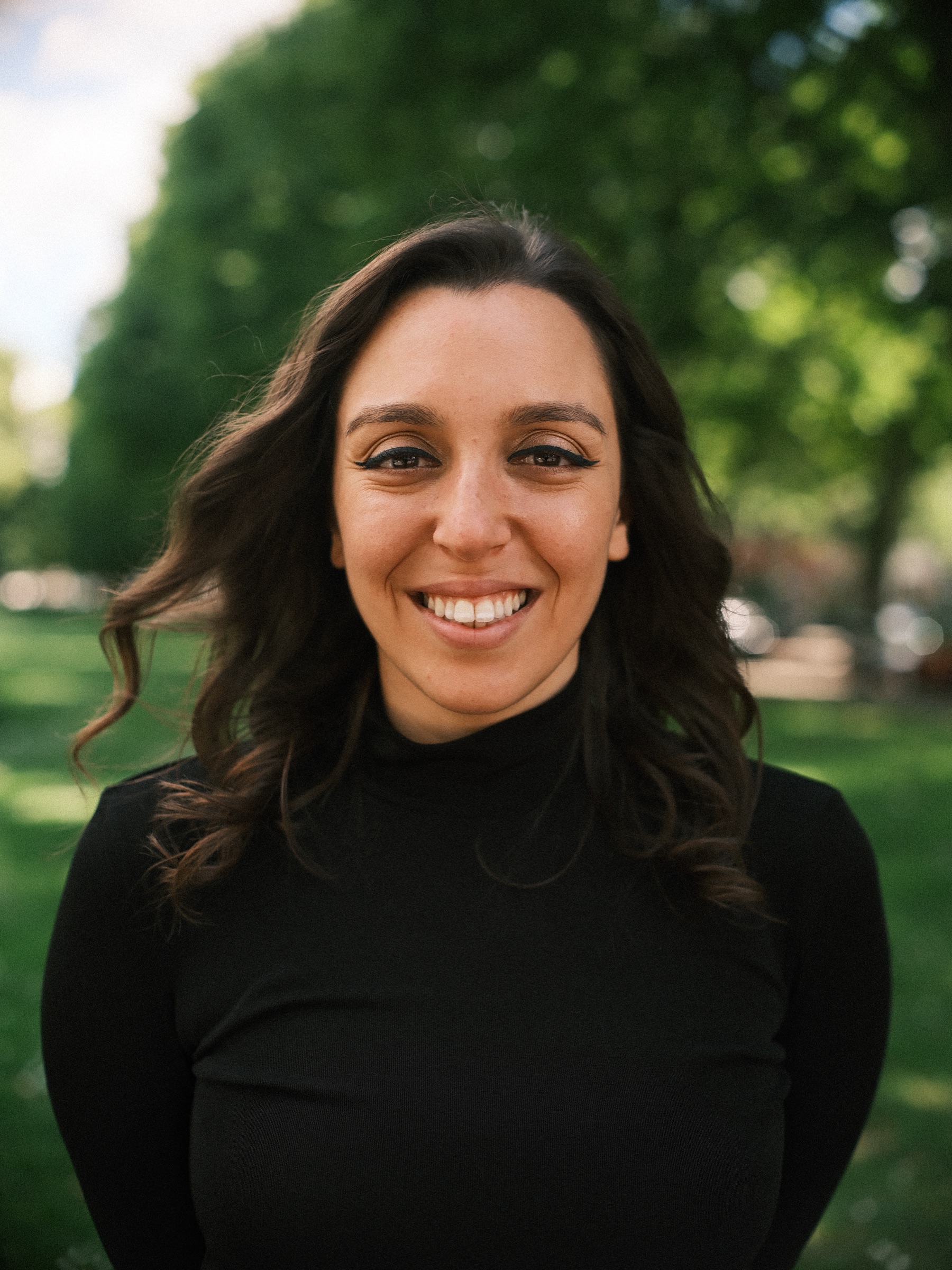 Head and shoulders portrait of her smiling in a black turtleneck, long dark curls, green trees softly blurred behind her