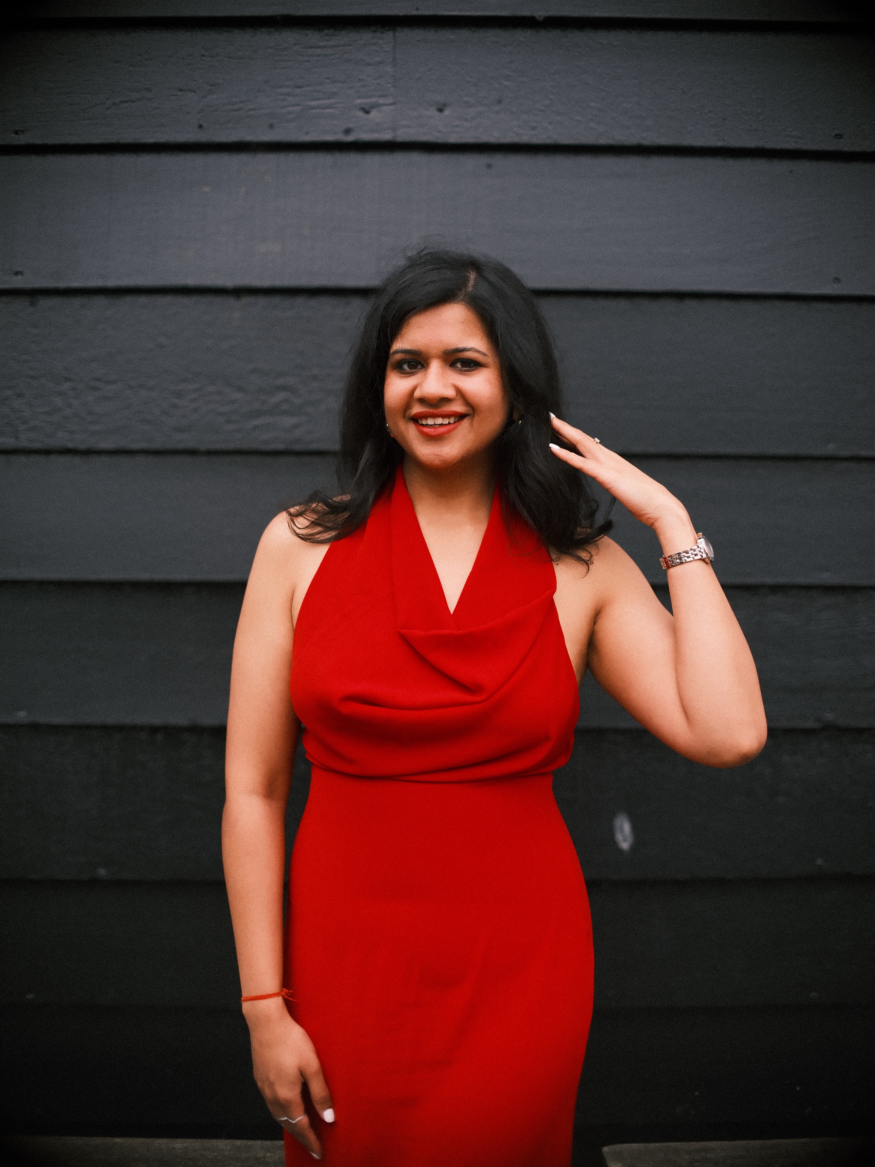 Portrait of a smiling woman in a red halter dress, one hand in her dark hair, against a dark wooden wall