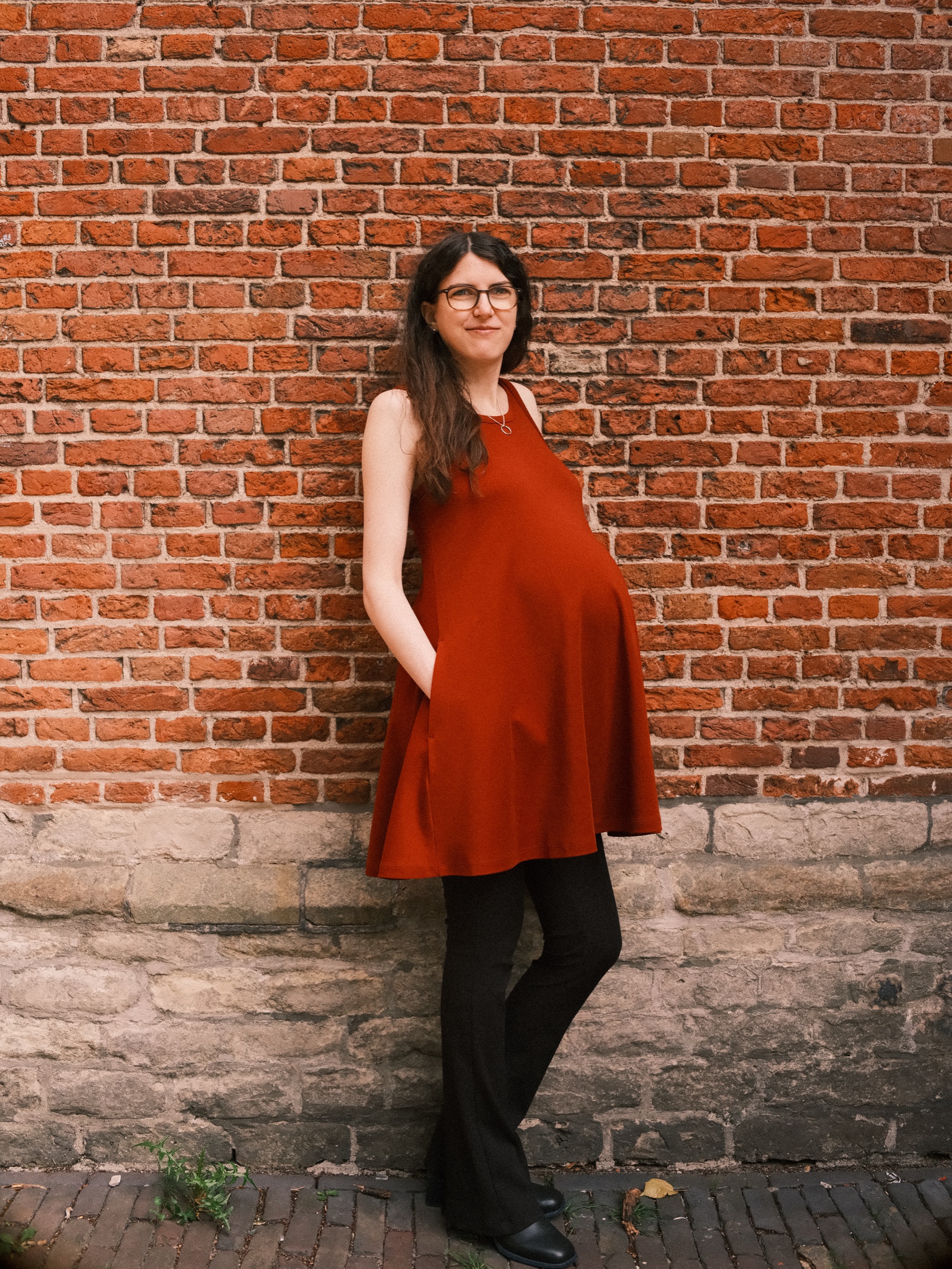 Pregnant woman leaning on a red brick wall above an old stone base, one hand in her pocket and black trousers under her dress