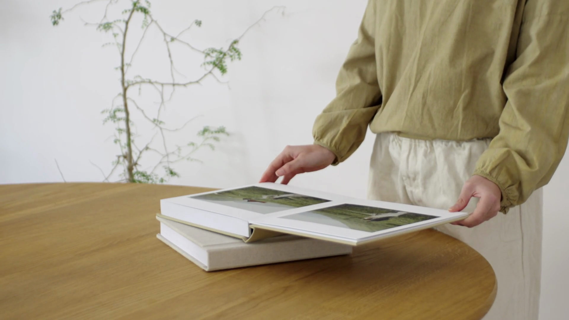 Hands opening a lay-flat photobook with landscape photos on a round wooden table, in front of a white wall with a thin green branch