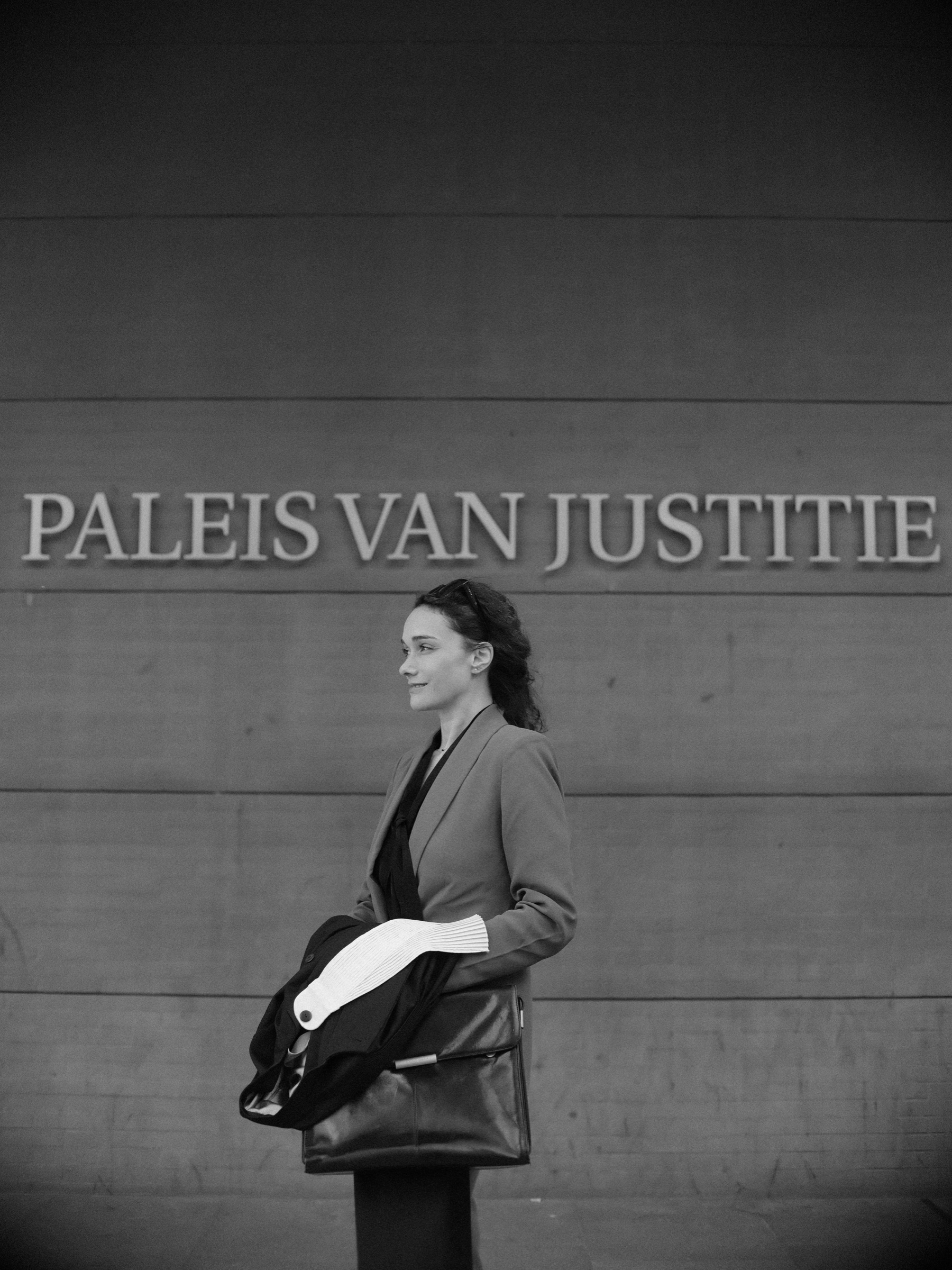 Advocaat in profiel kijkt omhoog onder de naam van het Paleis van Justitie, met haar opgevouwen toga en leren tas in haar armen