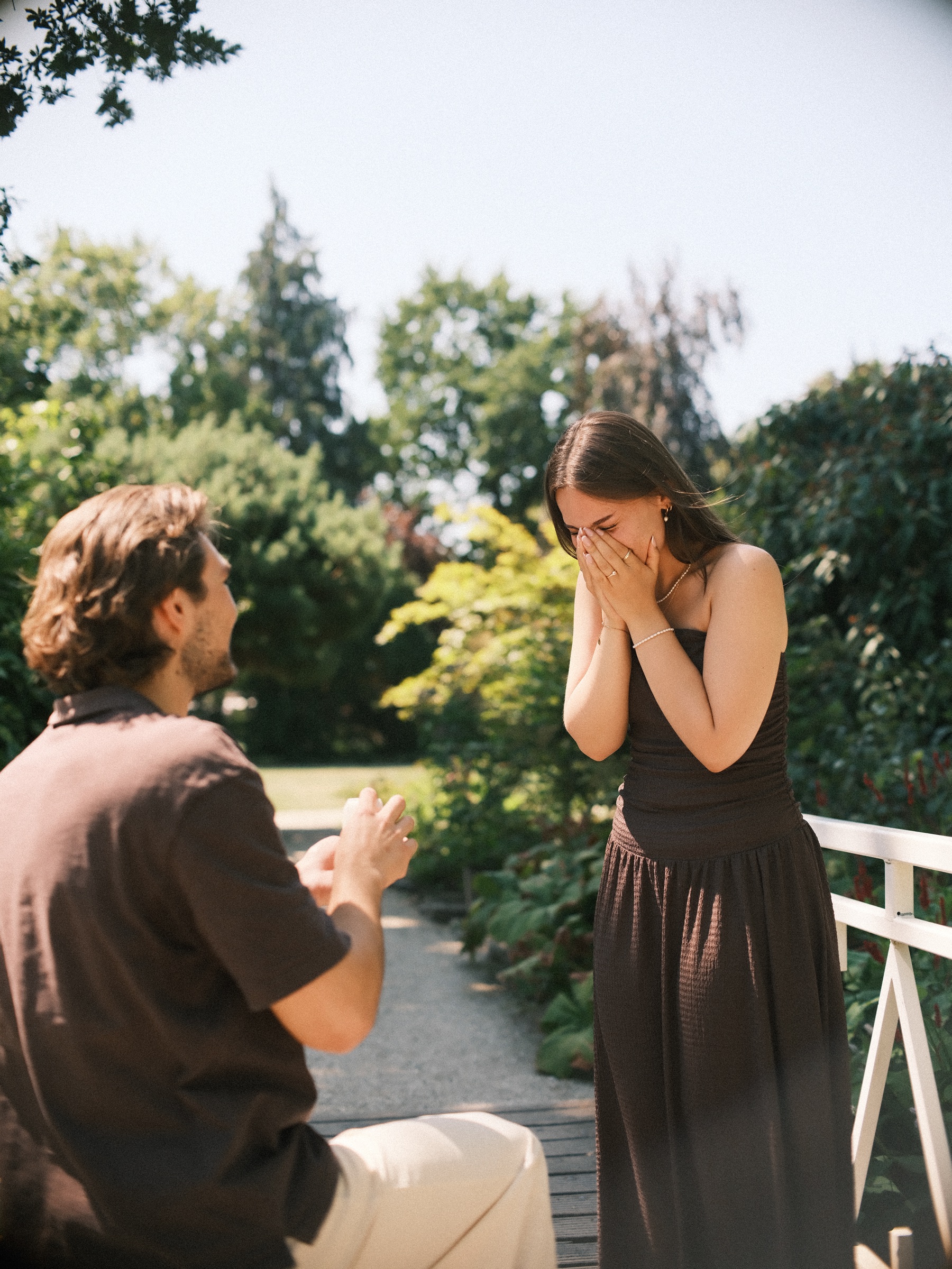 She covers her face with both hands as he kneels with the ring on a white wooden bridge, the surprise proposal at Trompenburg