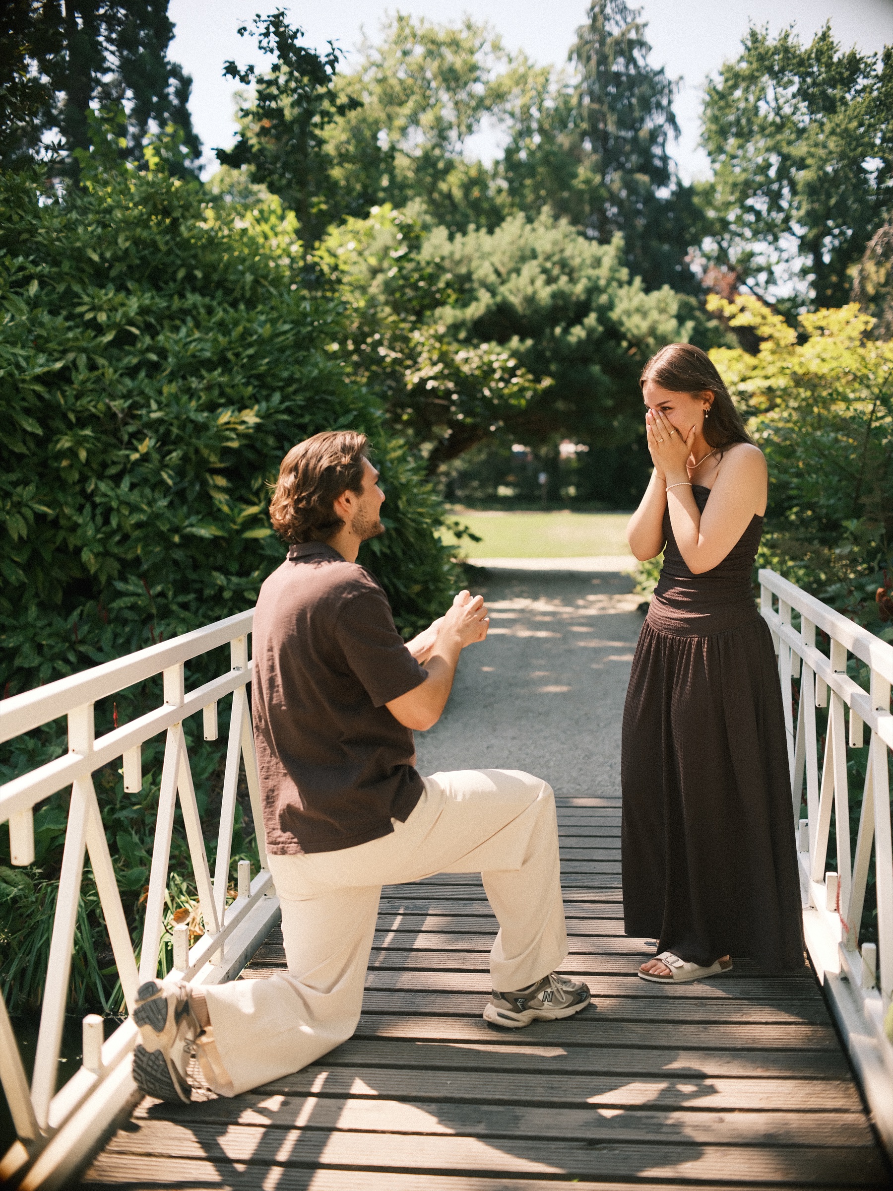 Man on one knee on a white wooden bridge while she covers her face with both hands, a surprise proposal in the Trompenburg gardens