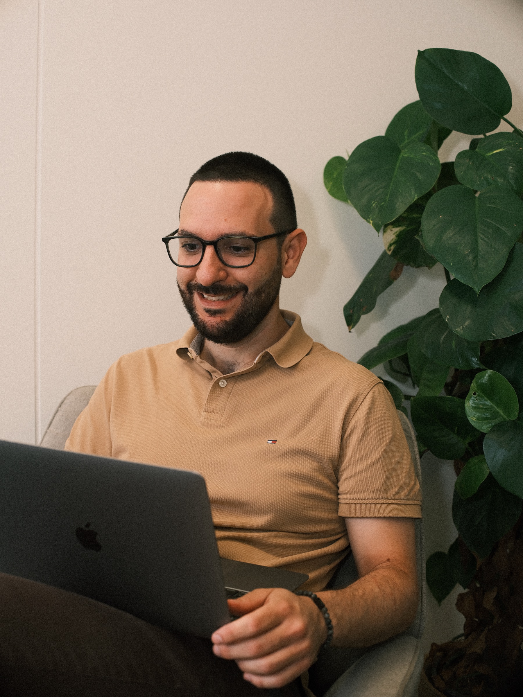 Man with dark glasses frames and a beard laughing over his laptop beside a large monstera, wearing a tan polo shirt