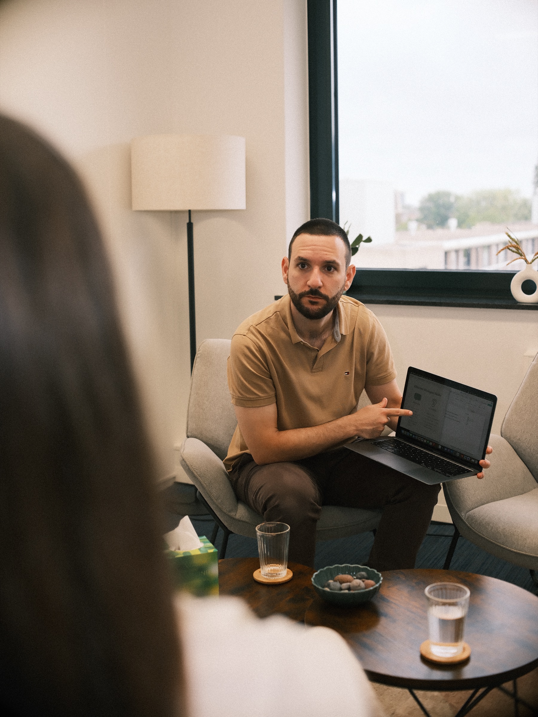 Man in a tan polo explaining something with a laptop on his knee, seen past the long dark hair of a woman in the foreground