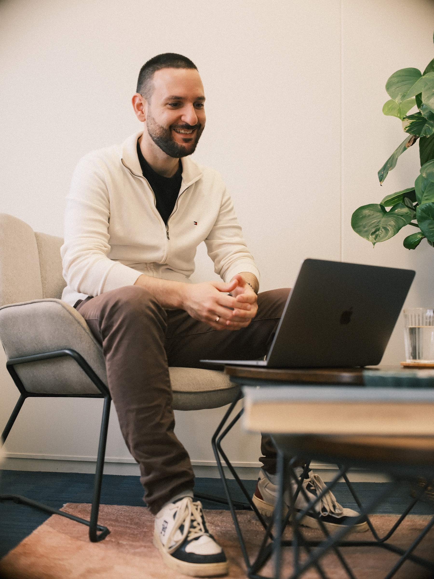 Psychologist in a cream zip jacket laughing in an armchair, hands clasped above a laptop on a small round wooden table