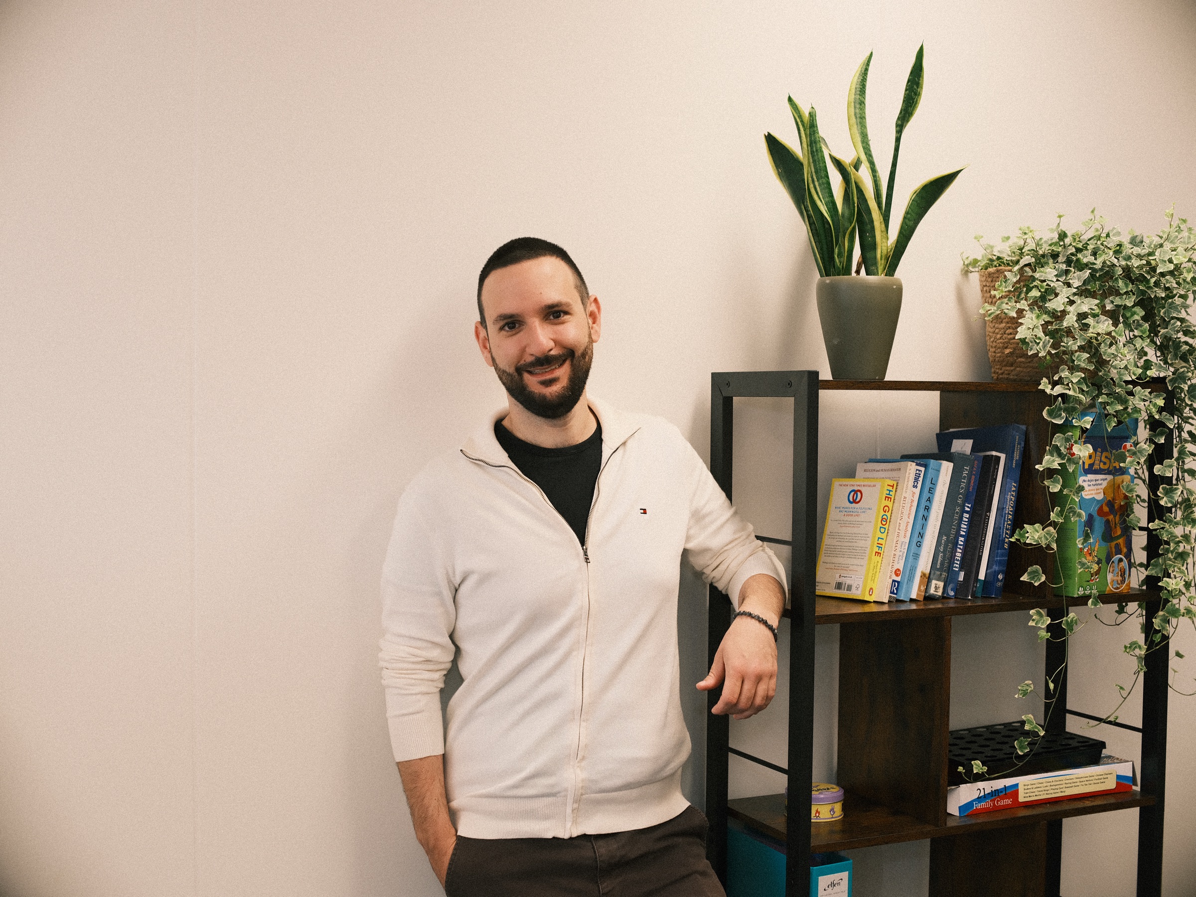 Smiling man in a cream zip jacket resting a hand on a dark bookcase with books, a snake plant and ivy in the therapy practice