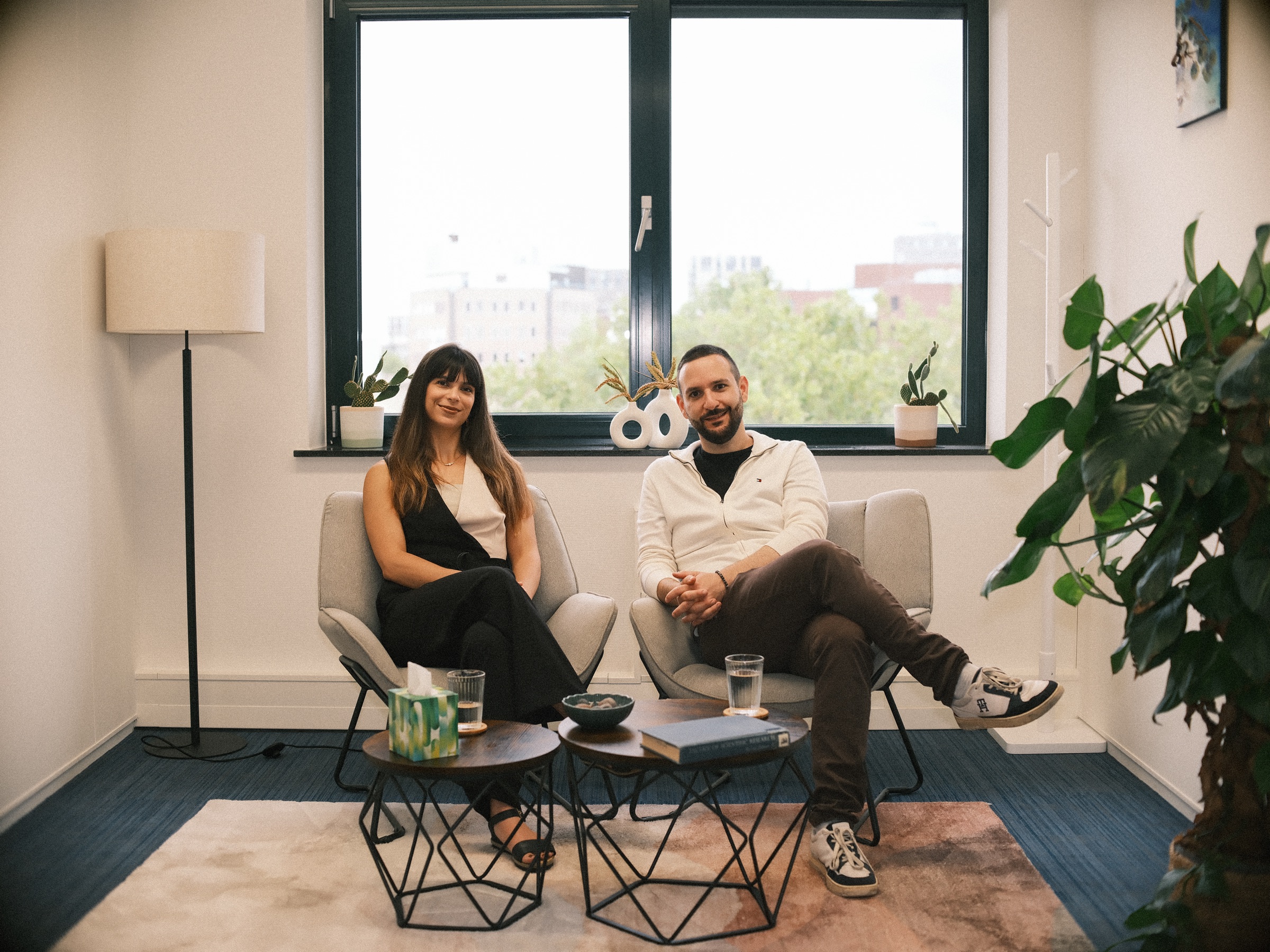Two psychologists of Therapy Care in gray armchairs by the big window of their therapy practice in Leiden, a monstera in front
