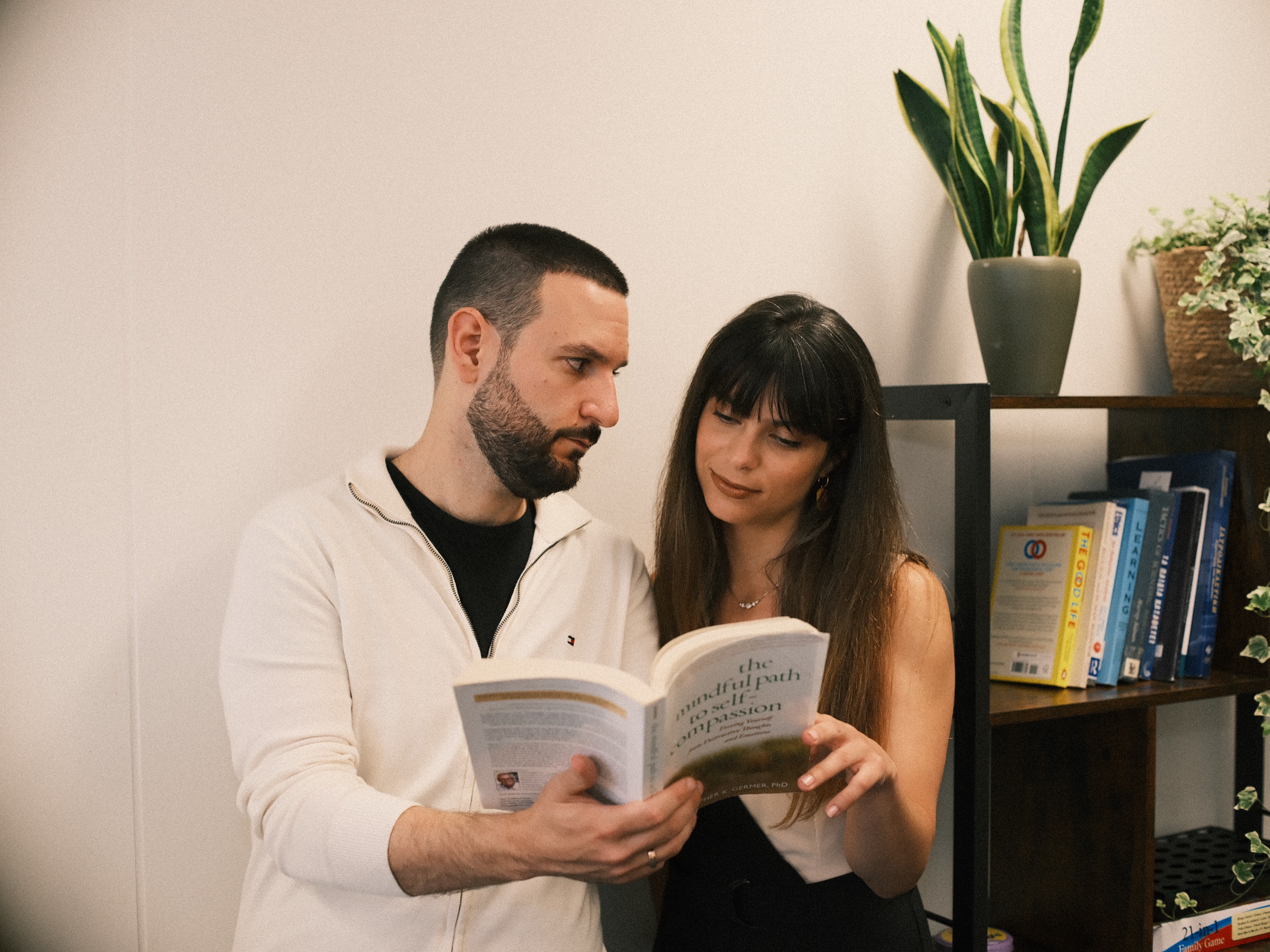 Two psychologists reading a book together by a bookcase with a snake plant on top, a candid moment from their branding shoot