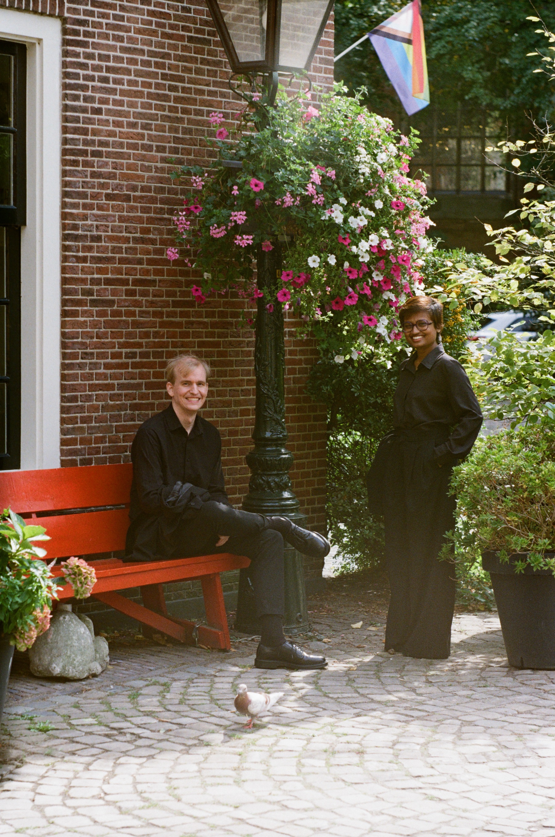 Man in black on a red bench below a lamppost with a hanging basket of pink flowers, his partner standing beside the plants