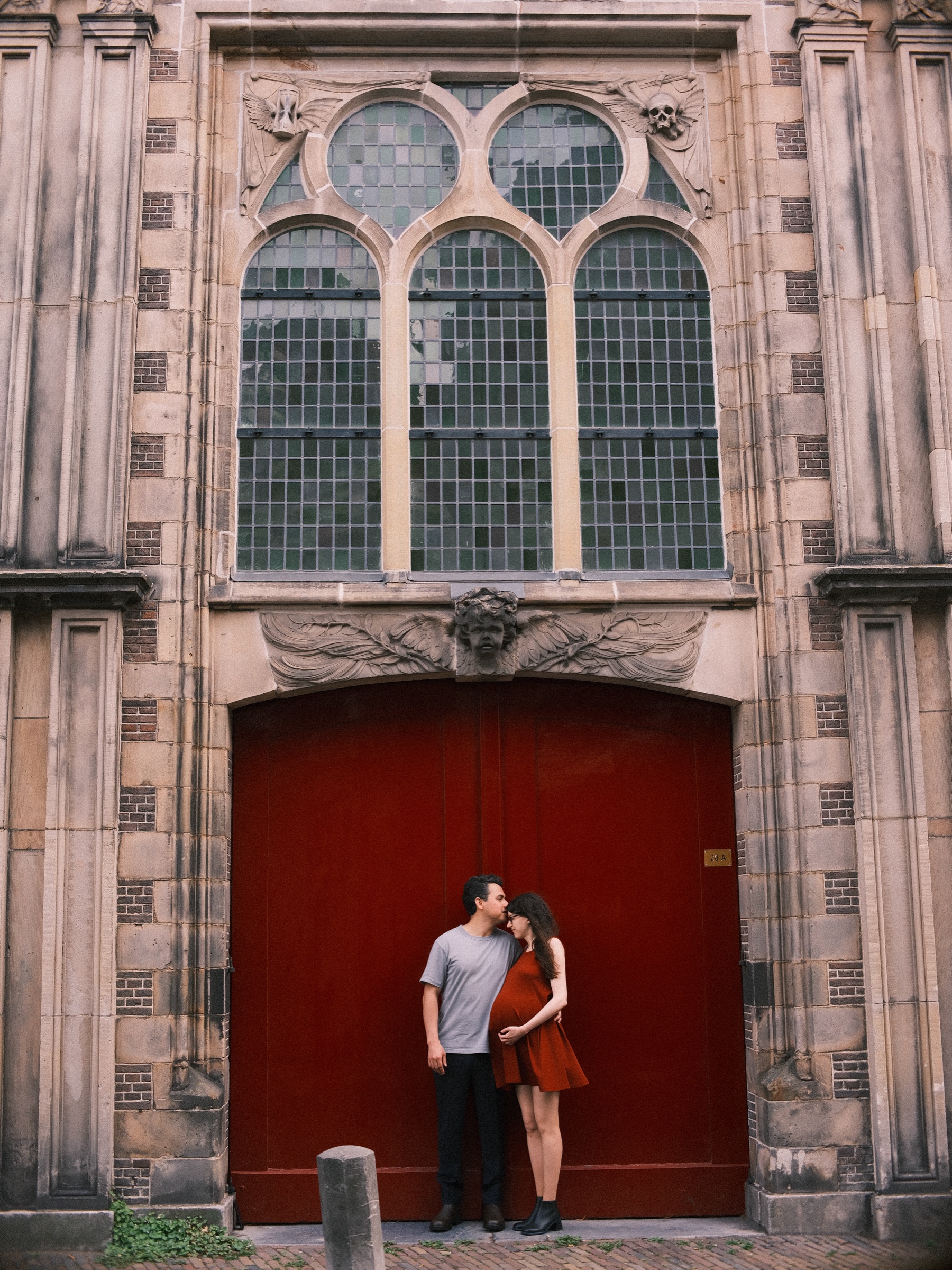 Tall stone facade with three arched windows and carved winged skulls above a deep red door, the expecting couple small at its foot as he kisses her hair