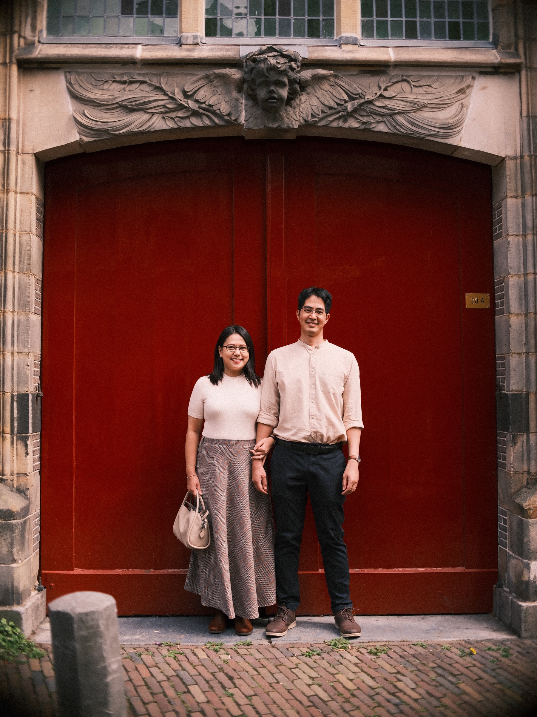 Couple standing side by side before red double doors under a carved stone face with wings, the whole old doorway in the frame