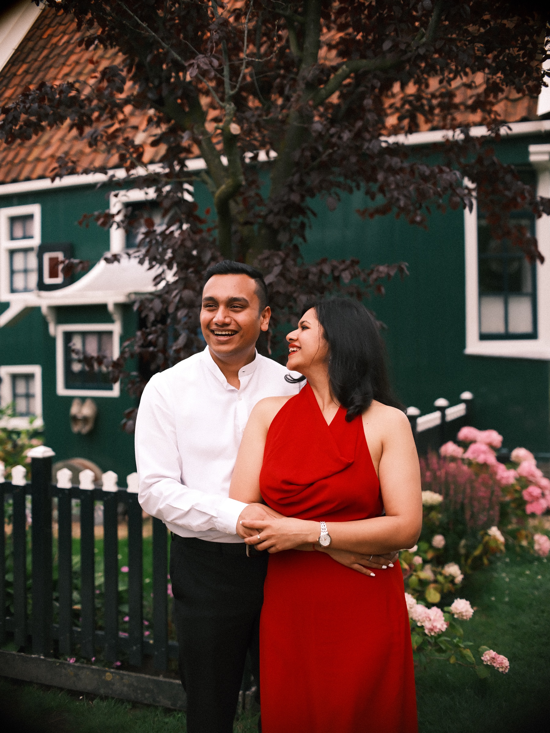 Laughing couple in front of a green wooden house with a red-leaved tree, she in a red halter dress and he in a white shirt