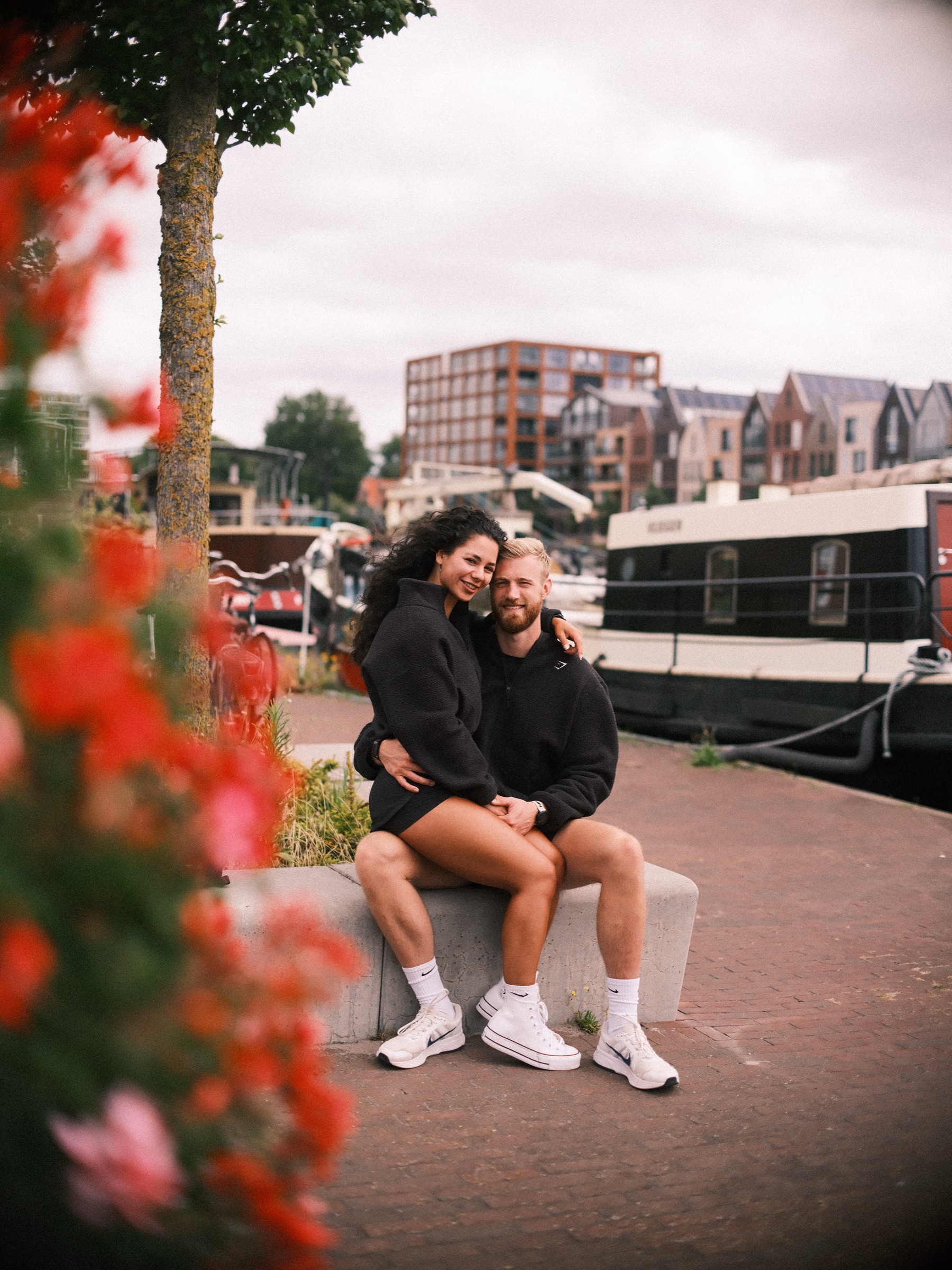 Rode geraniums vervagen voor het stel op een bankje bij een woonboot, met nieuwe bakstenen appartementen erachter