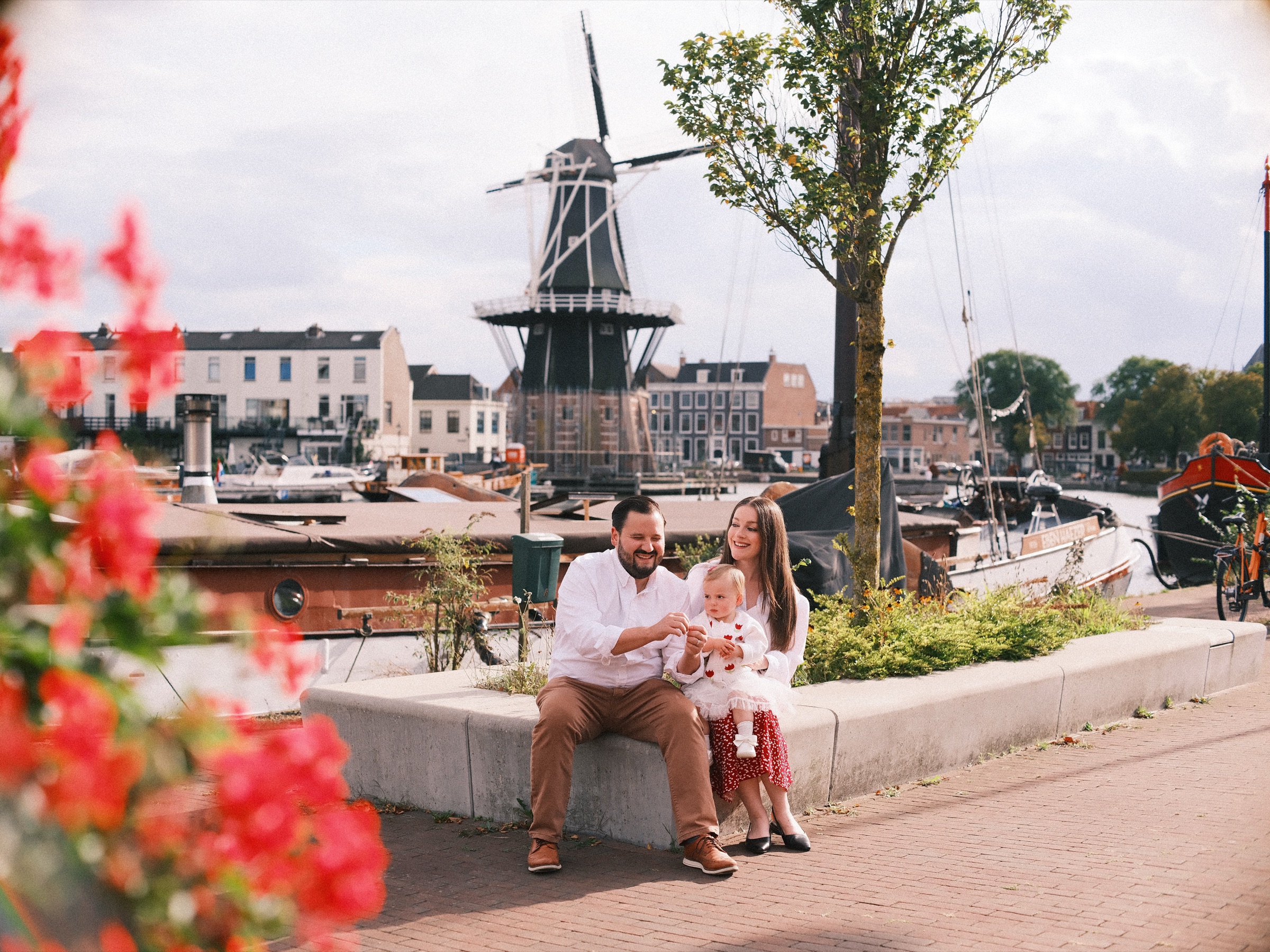 Rode geraniums wazig op de voorgrond, een jong gezin op een betonnen plantenbak met molen De Adriaan en boten erachter