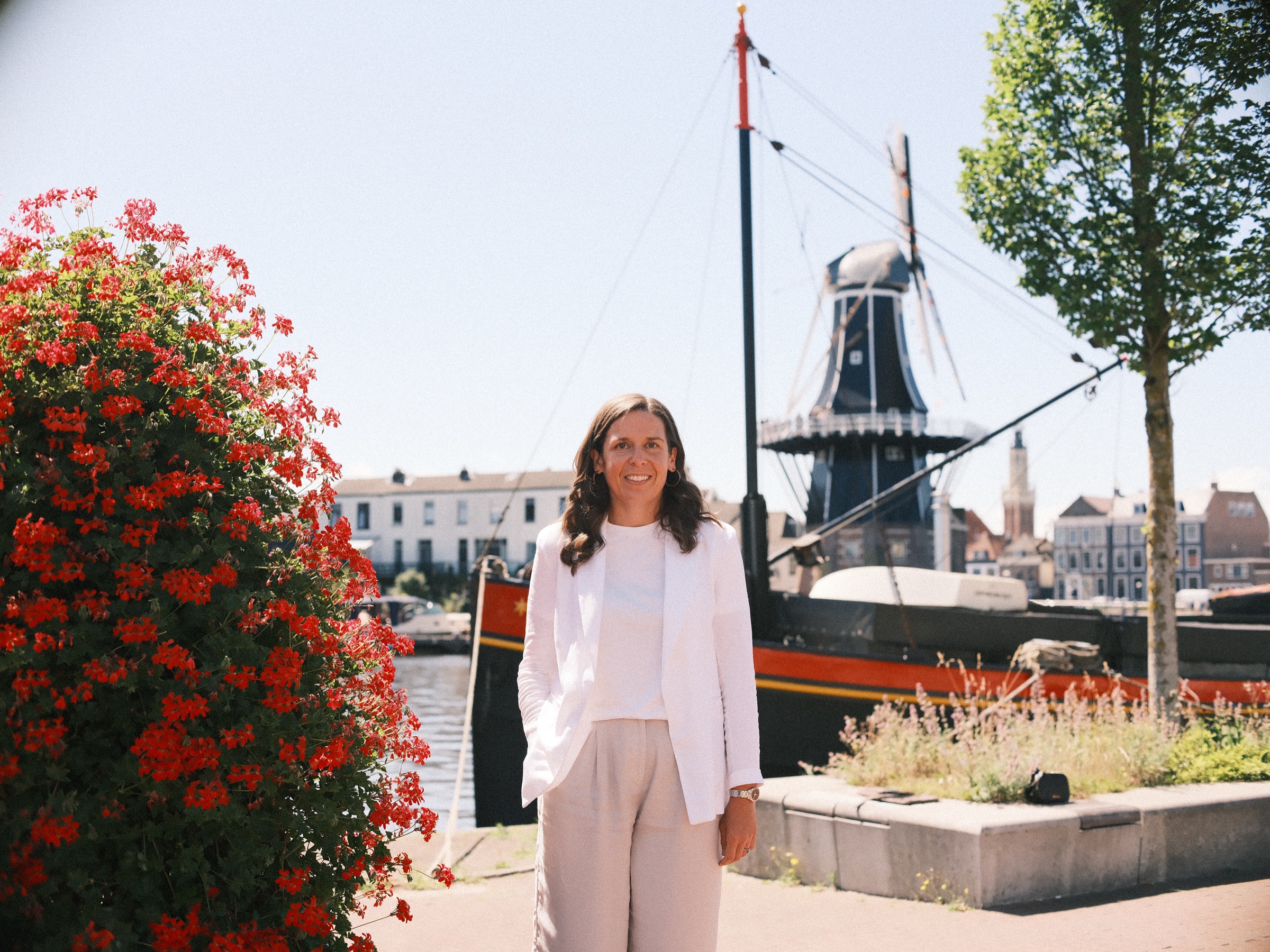 Vrouw in witte blazer en wijde broek bij een toren van rode geraniums, met molen De Adriaan en een oud schip erachter in Haarlem