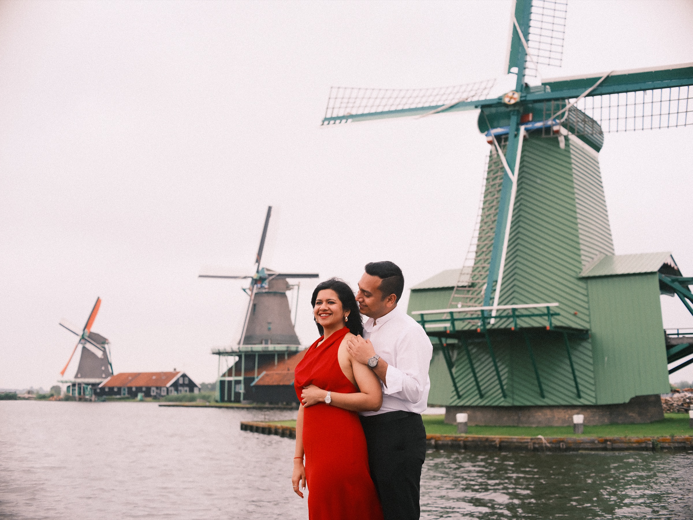 Woman in a red halter dress smiling as her partner holds her by the water, the windmills of the Zaanse Schans along the Zaan