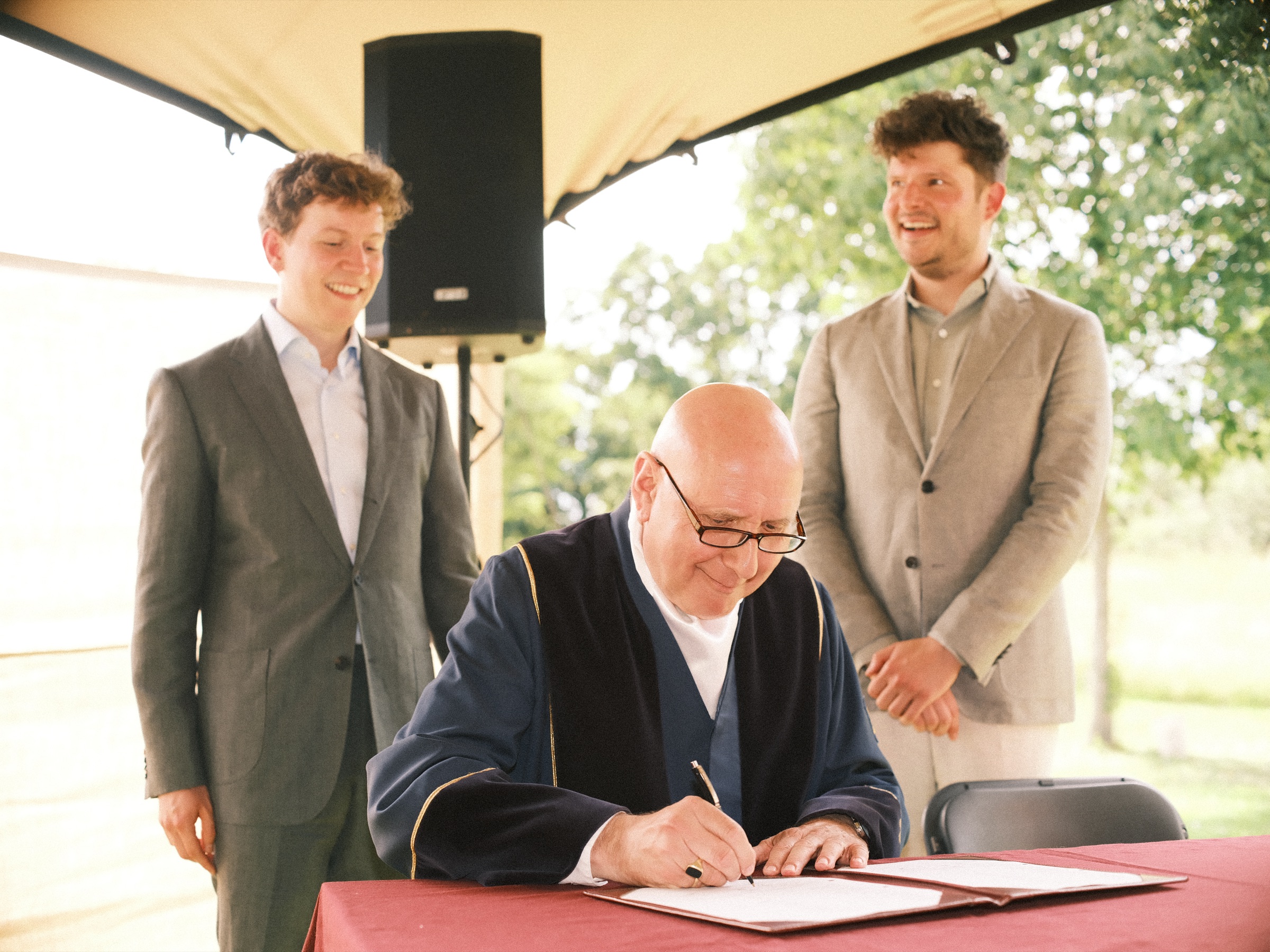 Registrar in a black robe signing the papers while both grooms stand beside him laughing, at their wedding in Utrecht