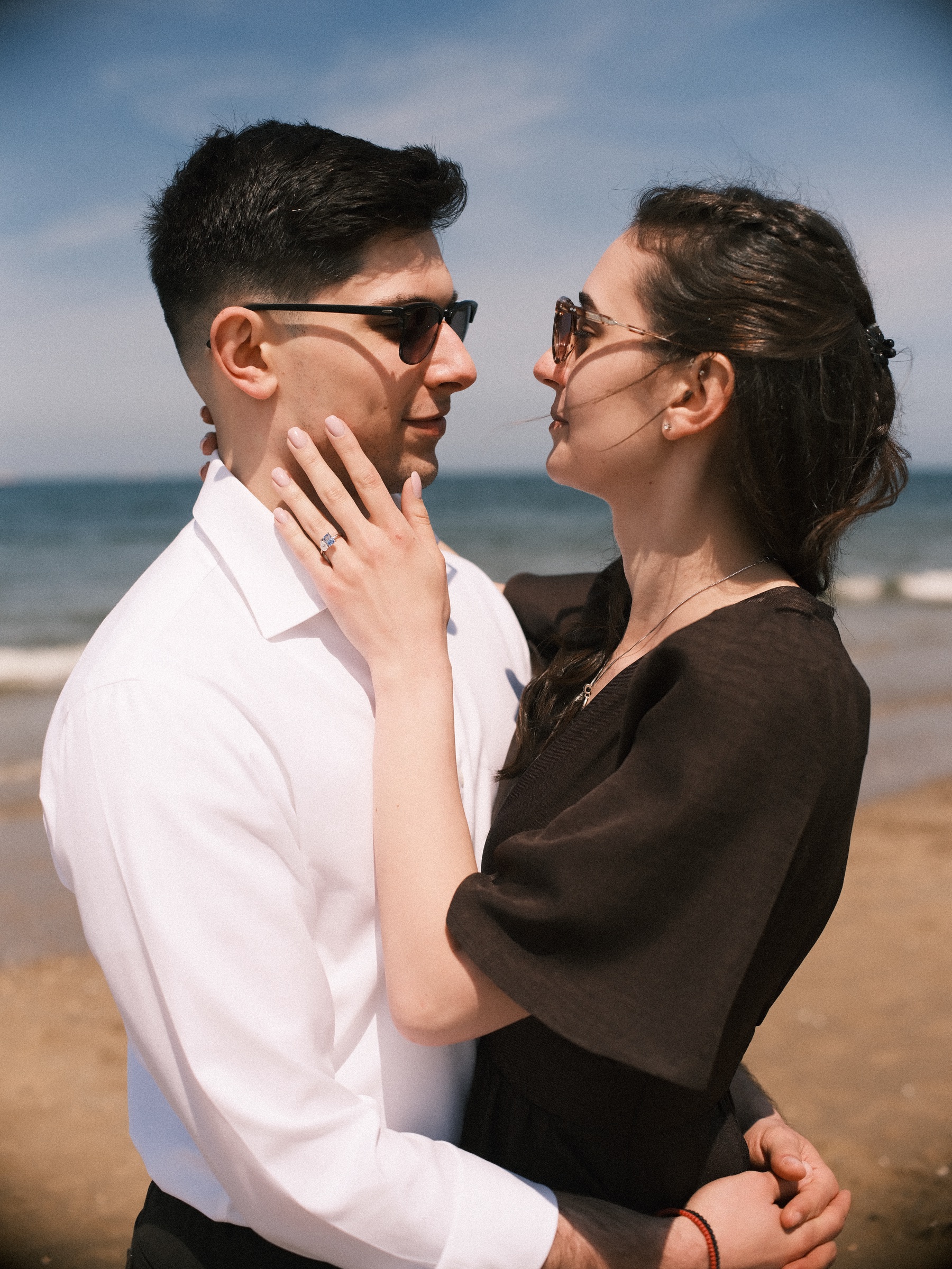 Her hand with the new blue ring resting on his cheek as they lean in close on the beach, both in sunglasses, the sea soft behind them
