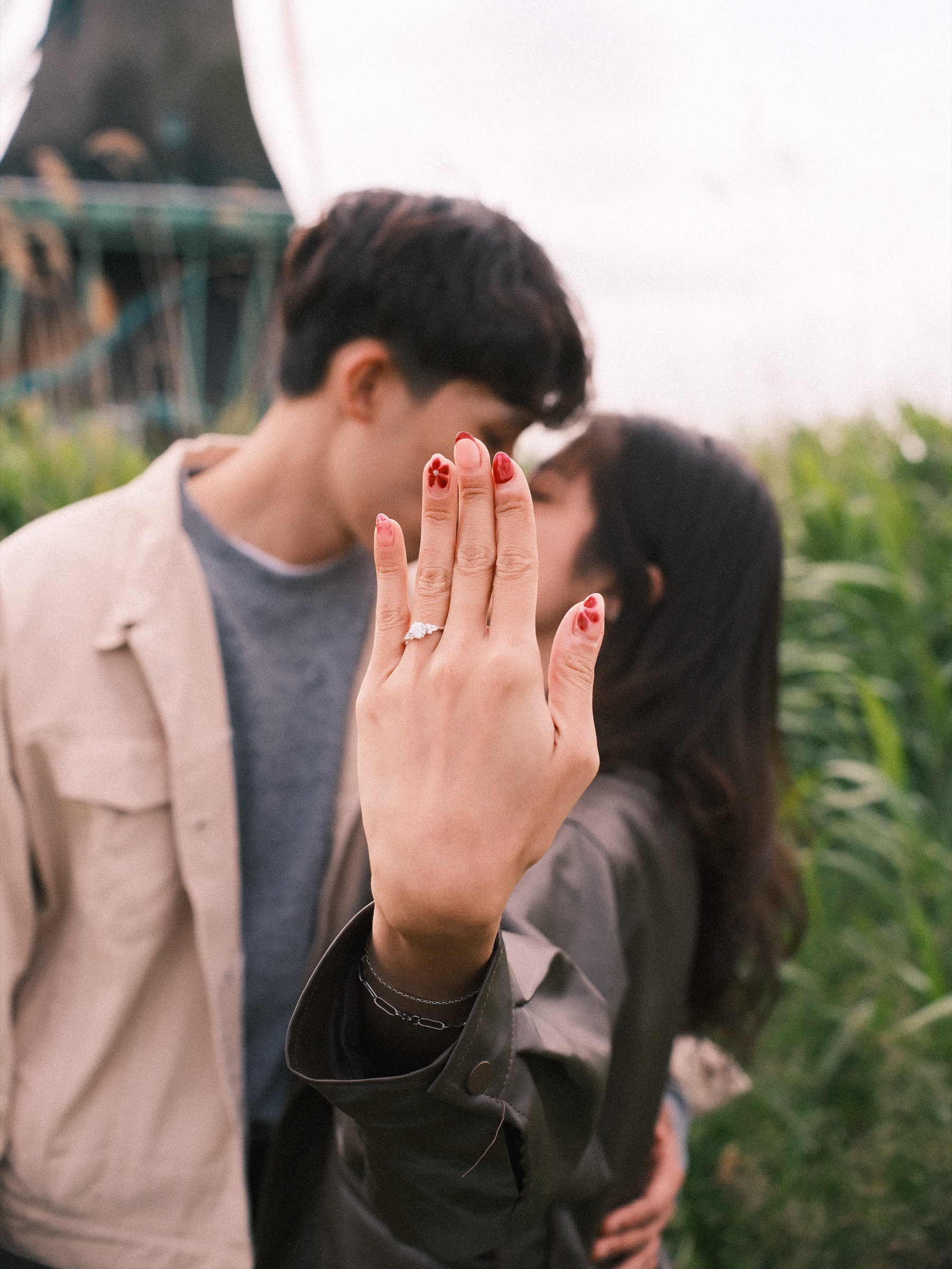 She holds her ring hand up in front of a kiss, her fiancé in a cream jacket half hidden behind it, tall green reeds around them