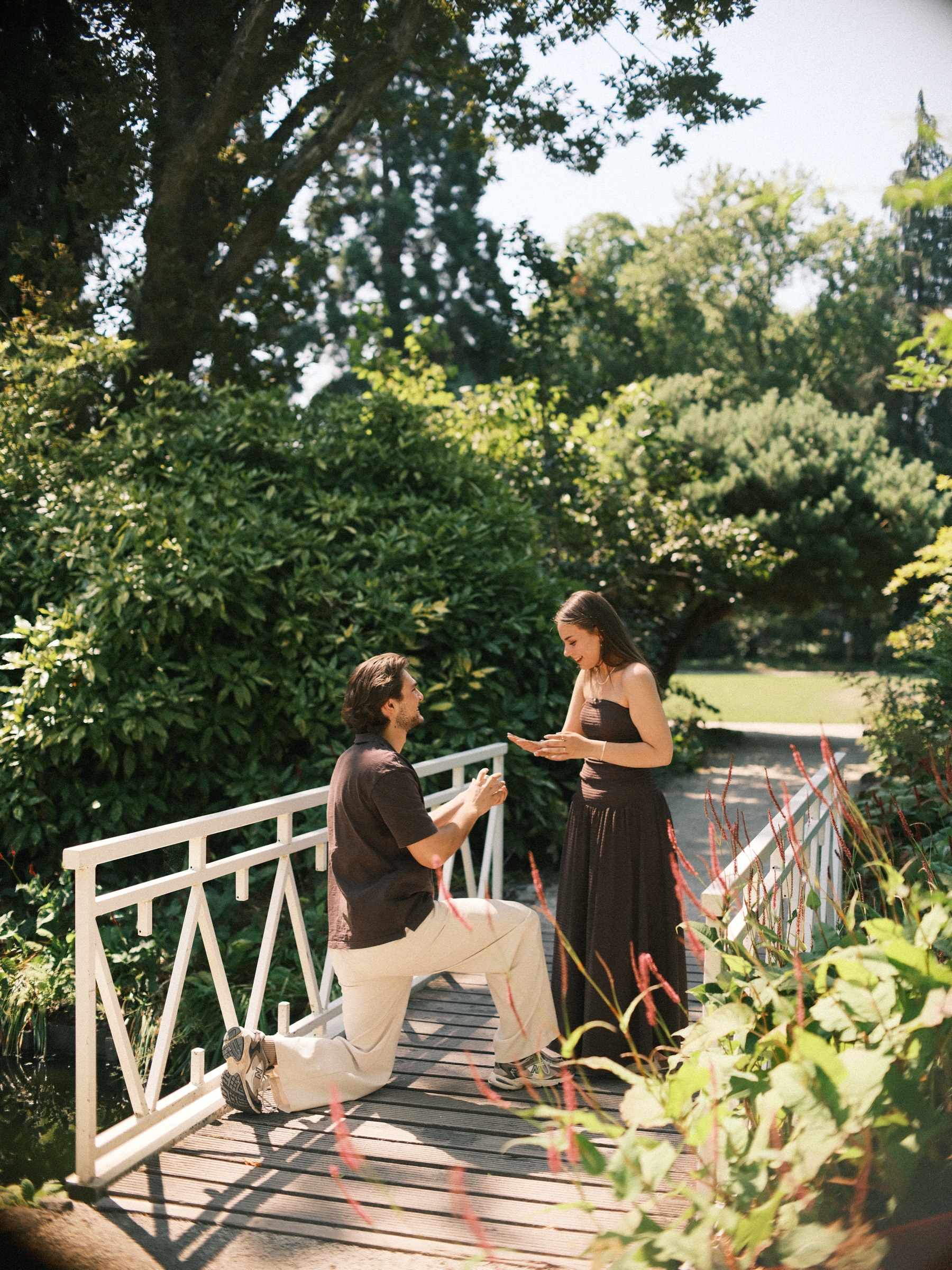 Kneeling on the planks of the white bridge, he puts the ring on her finger during the surprise proposal, red flower spikes in front