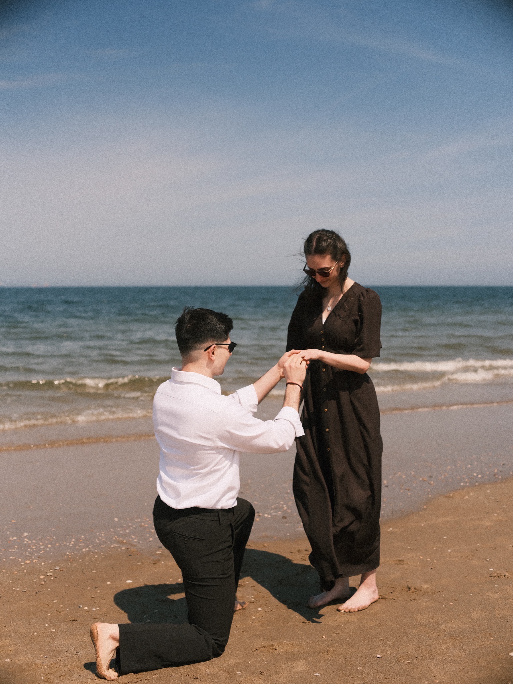 Still on one knee at the waterline, he holds her hand from below and slides the ring onto her finger, small waves rolling in behind them