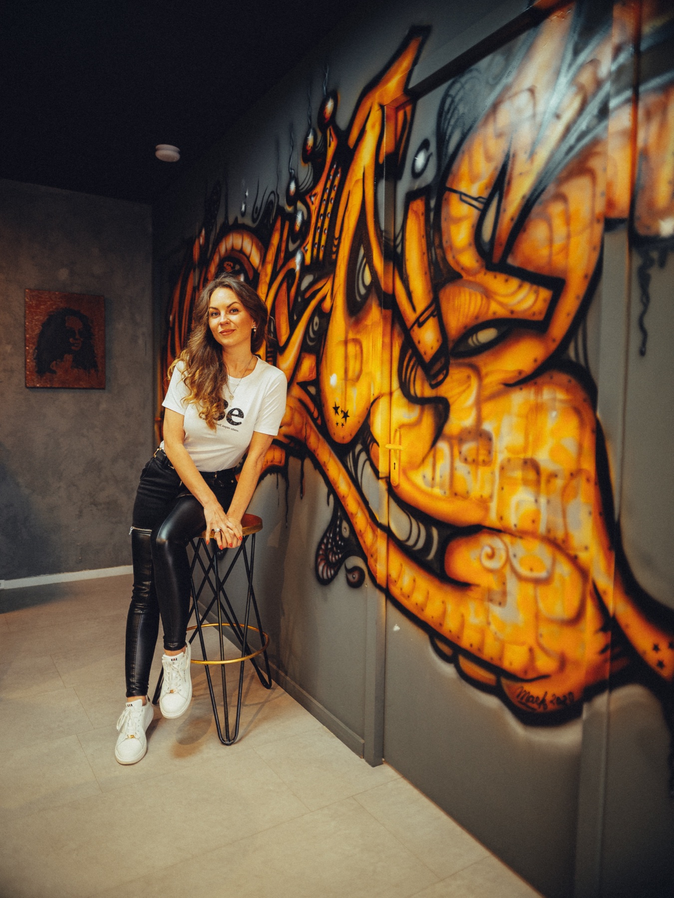 Eigenaar zit op een barkruk voor een oranje graffitimuur in haar beautystudio, lachend in een wit Be-shirt