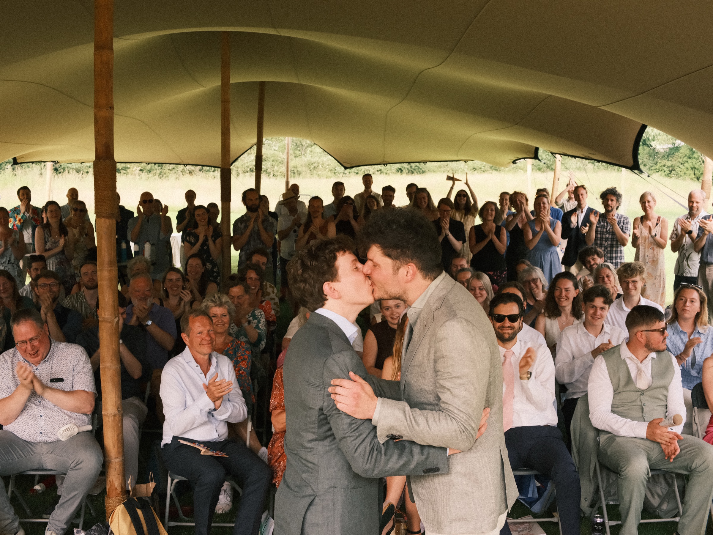 Two grooms kissing under a sand-colored tent as their guests clap and cheer at their same-sex wedding in Utrecht