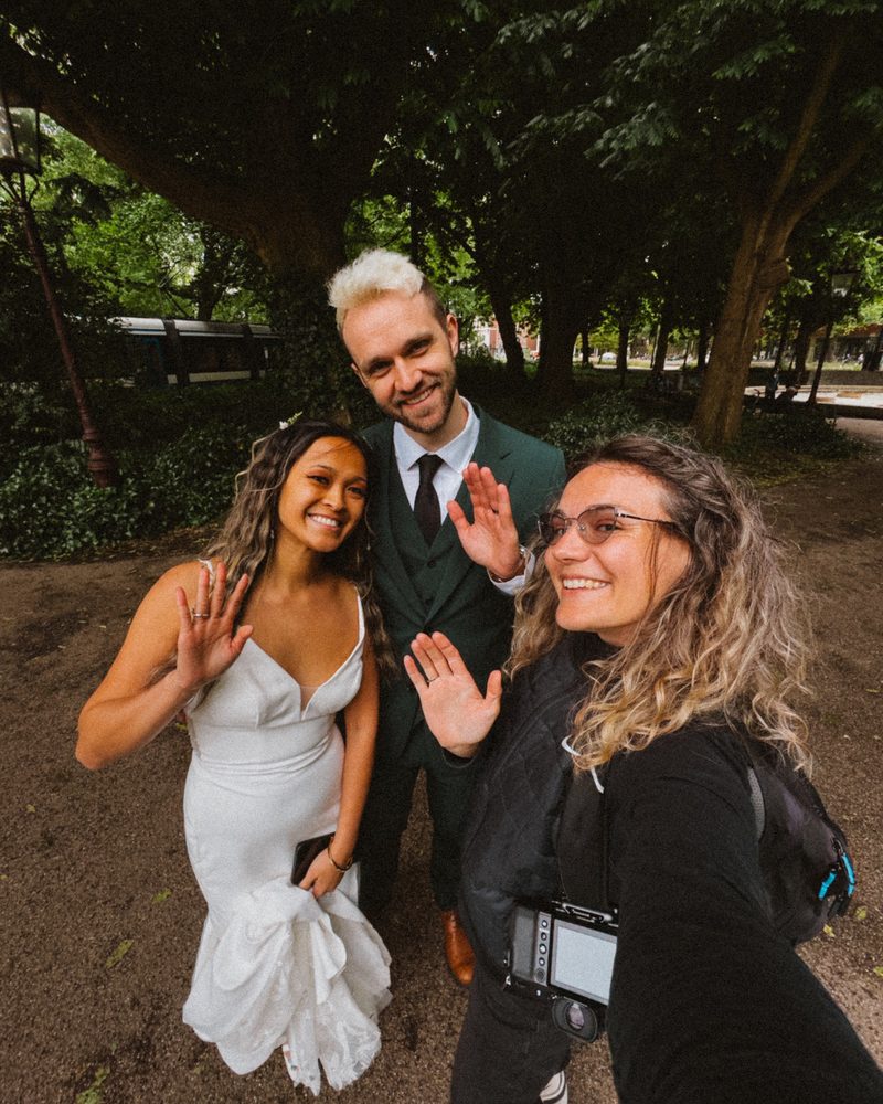 Bride in a white gown and groom in a dark green suit waving into a selfie under tall trees, a camera hanging on a strap in front