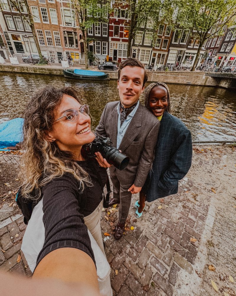 Wide selfie on an Amsterdam canal quay with a couple in tailored coats, a camera held in one hand and autumn leaves on the cobblestones