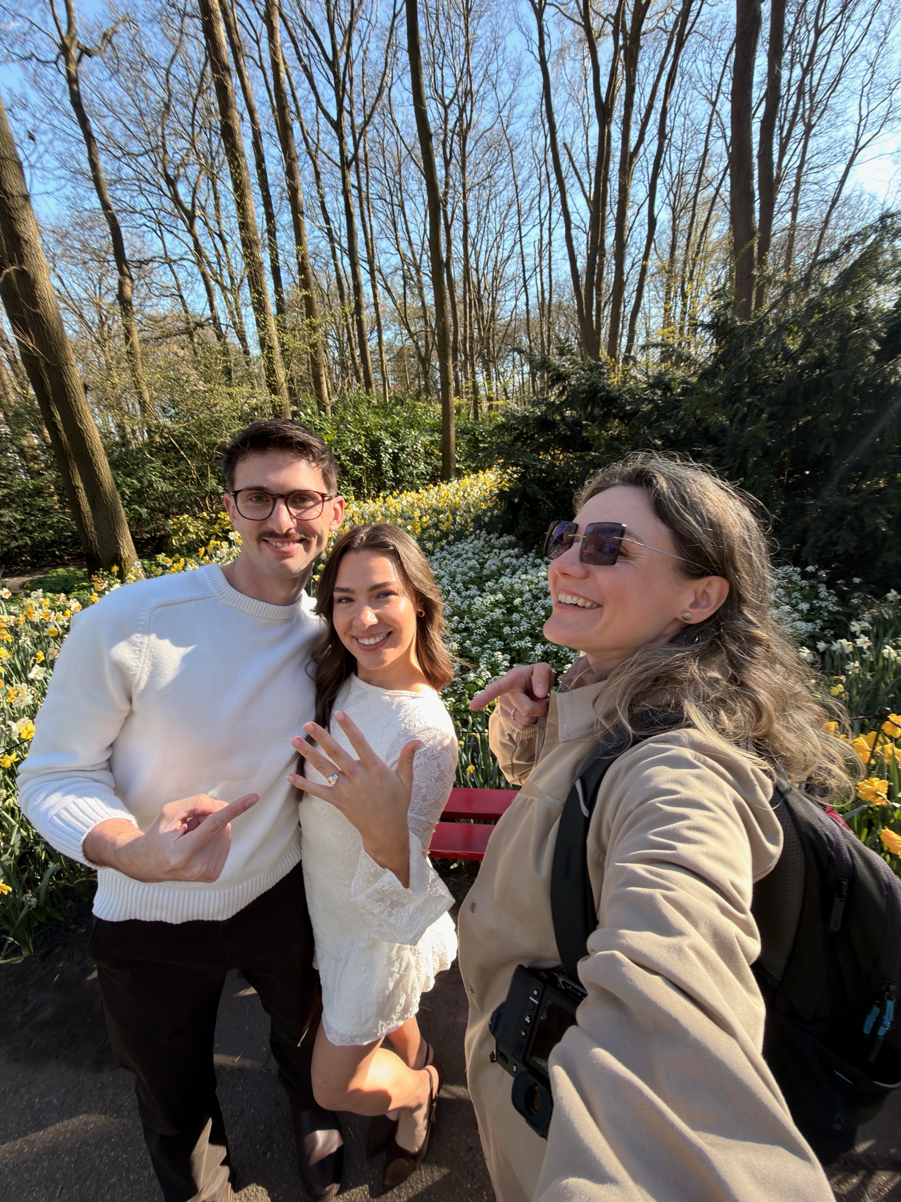 Selfie van een lachend stel en een vrouw met een analoge camera op een zonnig tuinpad, terwijl de jonge vrouw haar nieuwe ring laat zien