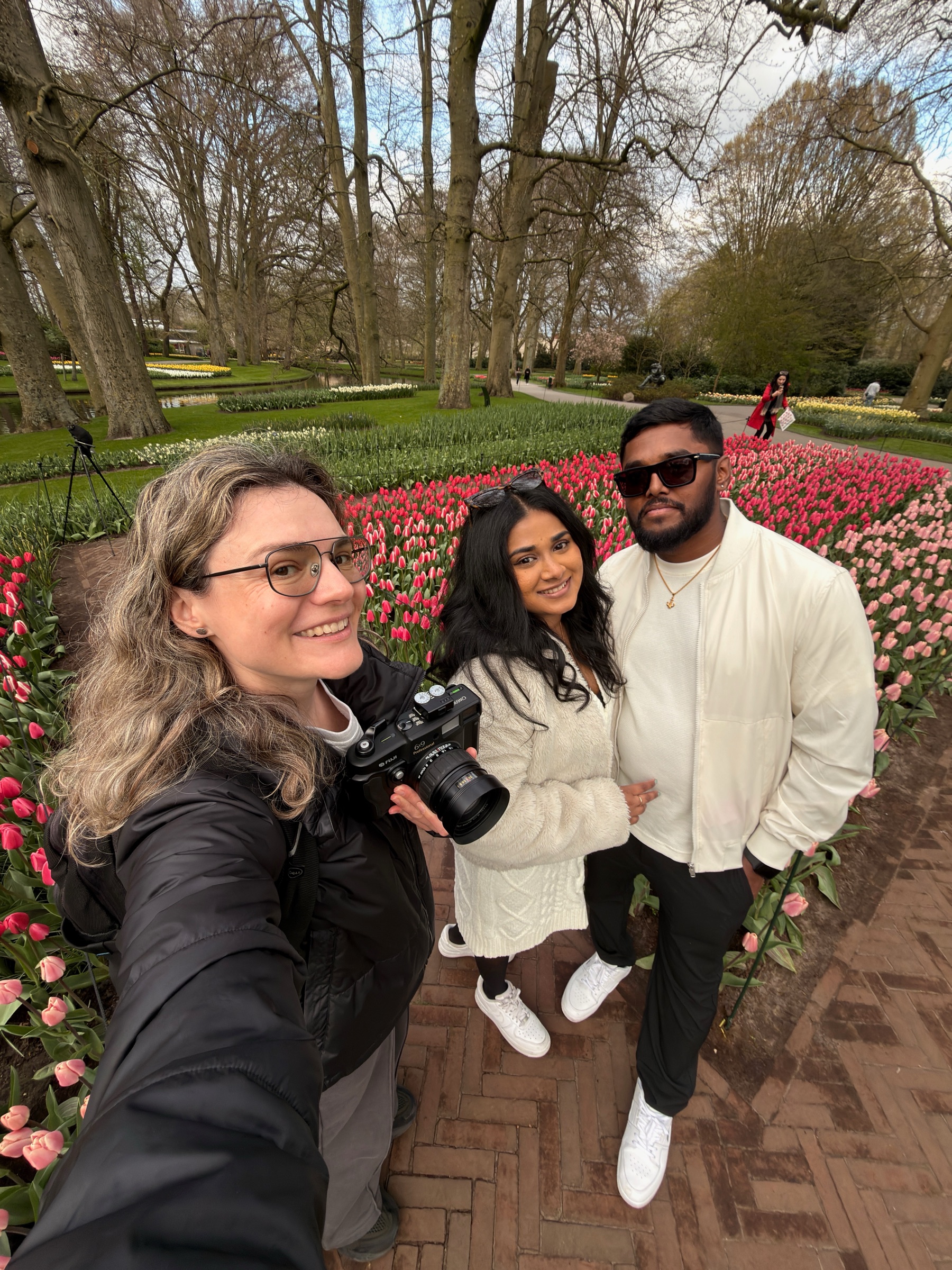 Selfie of a smiling couple in white with a woman holding a film camera, rows of pink tulips and a tripod behind them