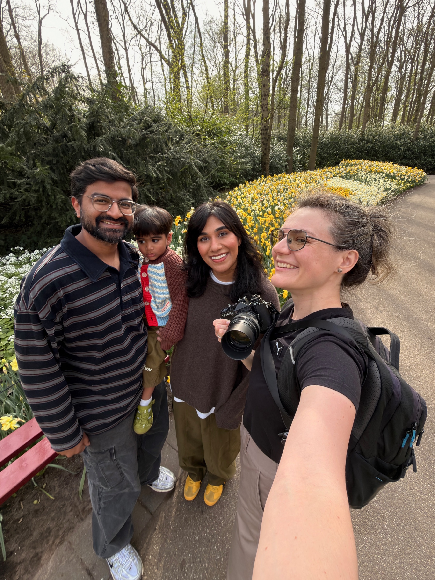 Selfie of a smiling family with a toddler in a red jacket and a woman holding a film camera, yellow daffodils behind them