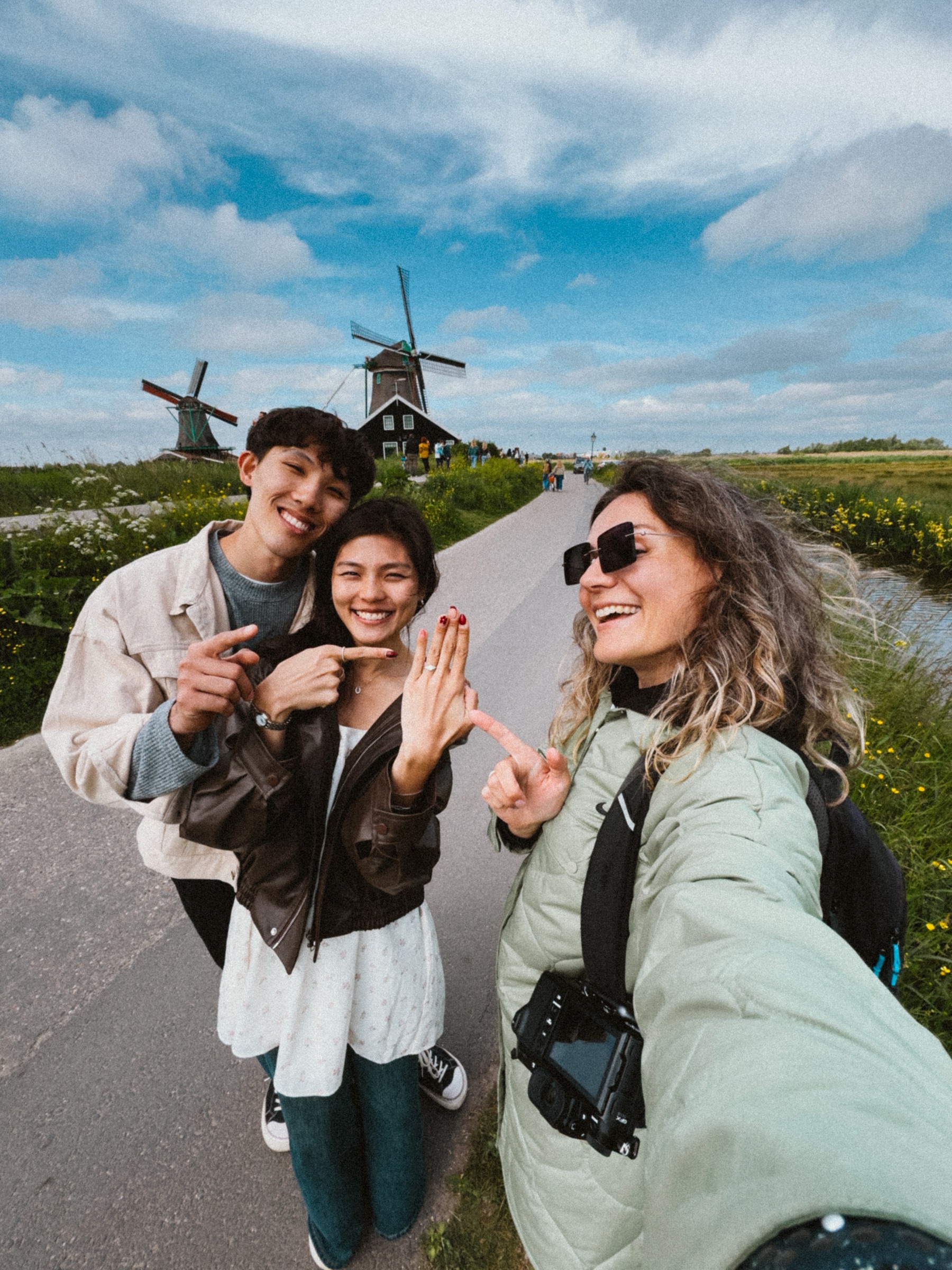 Selfie of the newly engaged couple pointing at her ring beside a smiling woman in sunglasses, windmills of the Zaanse Schans behind
