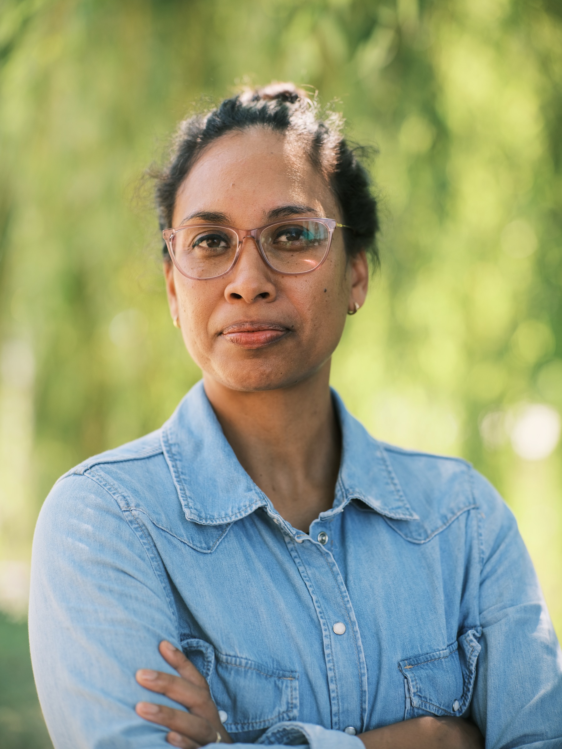 Portret van dichtbij: een vrouw met een rustige, serieuze blik, bril en knotje, armen gekruist in een spijkerblouse voor zachtgroene bladeren