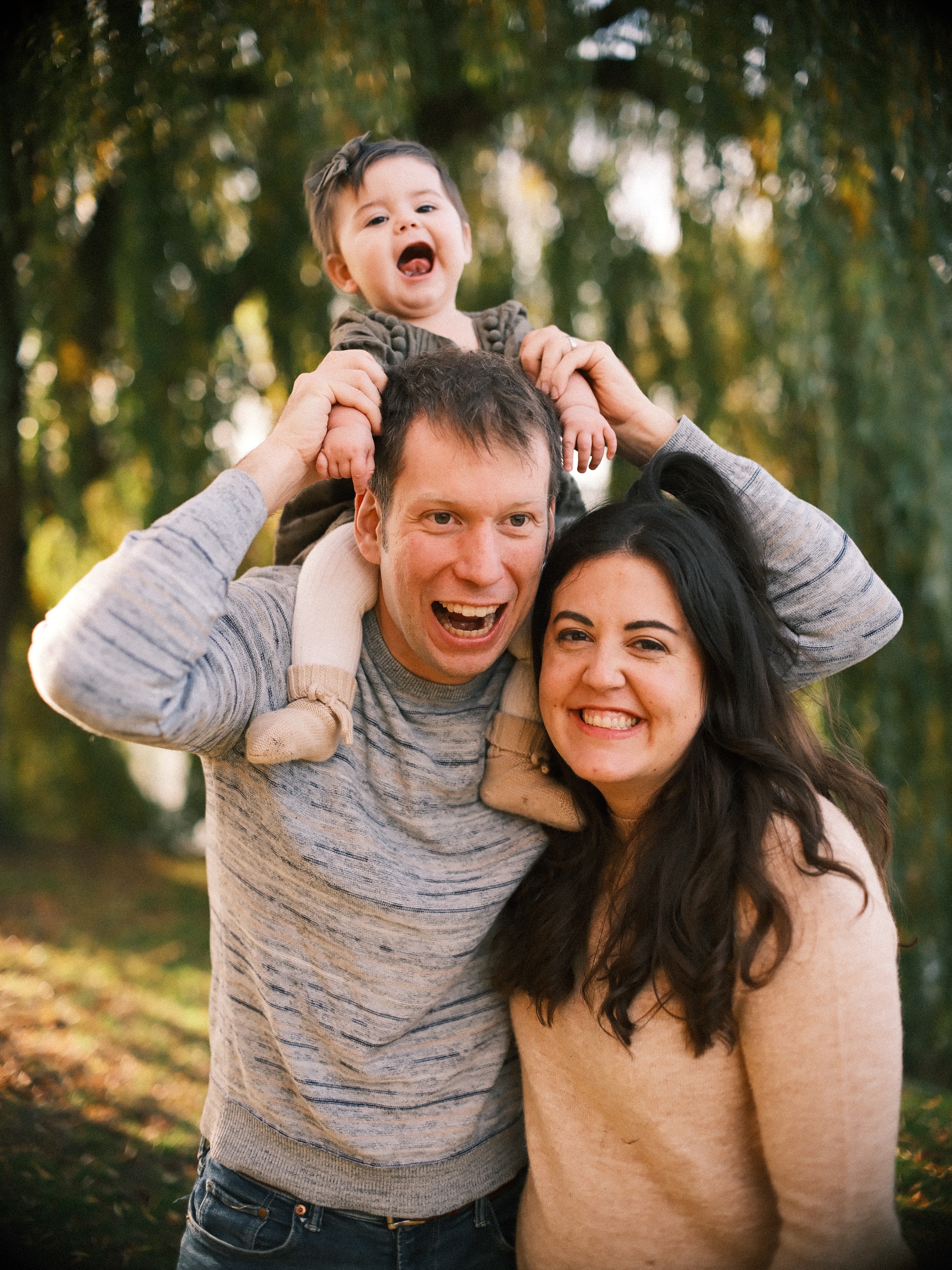 Father with his laughing baby daughter on his shoulders and the mother smiling beside them under a willow on an autumn day in Leiden